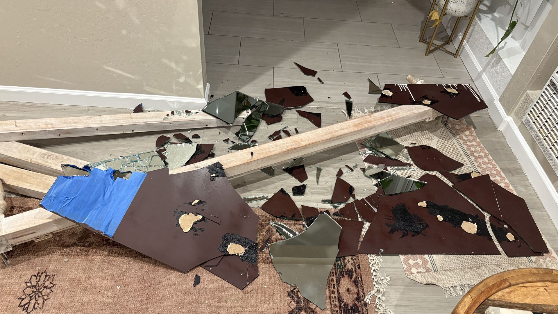 Shattered glass and brown laminate panels litter a light gray wood floor as two wooden beams span a doorway; blue painter's tape and a beige patterned rug lie nearby.