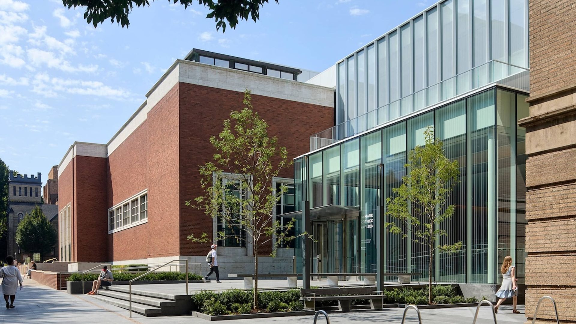 Outdoor view of the Mark Rothko Pavilion with modern glass facade and red brick walls, small trees planted around, people walking and sitting, under a blue sky with some clouds.