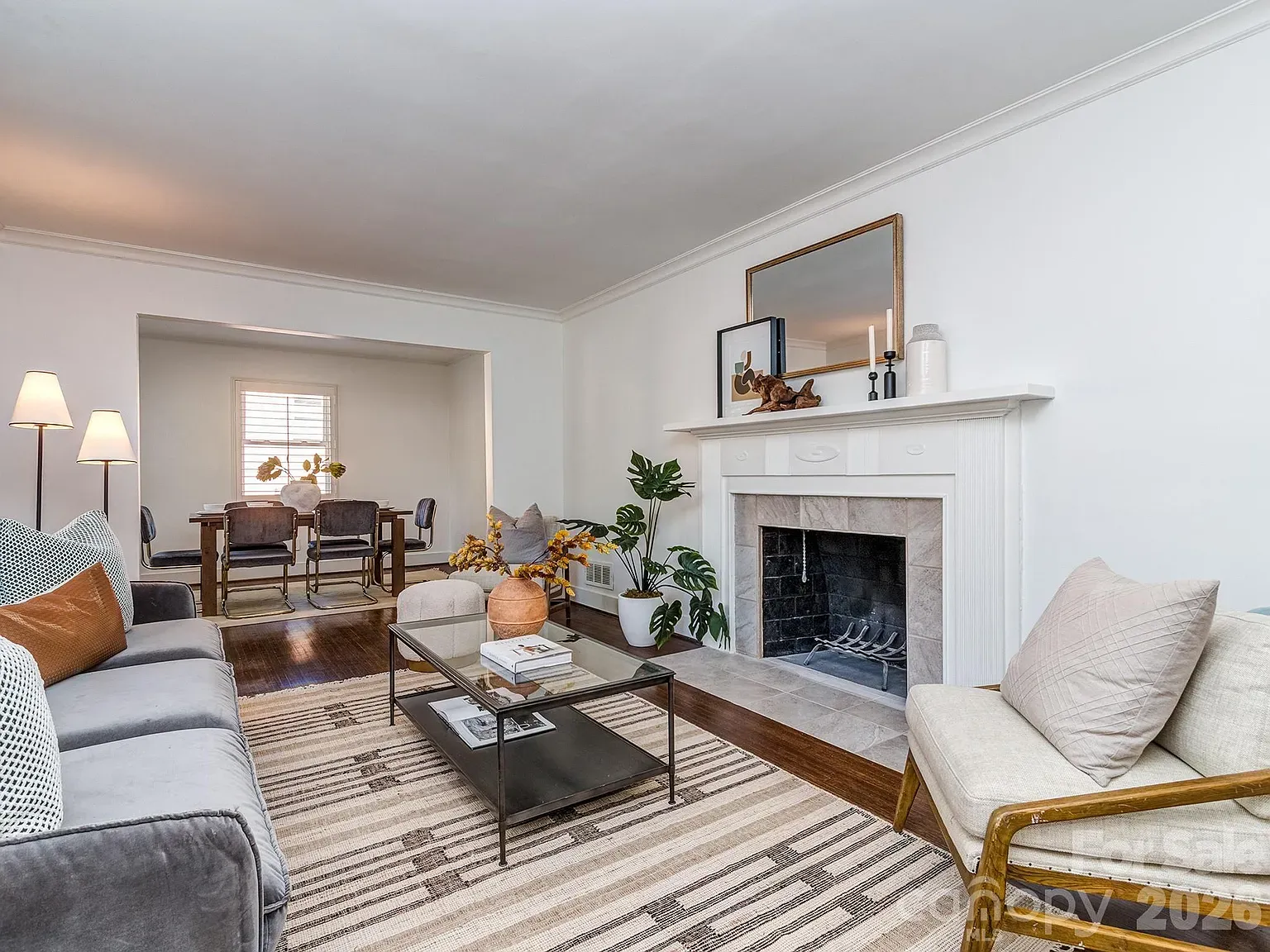 Bright living room with white walls and crown molding, a white mantel fireplace, gray sofa and cream chair, glass coffee table on a striped rug, potted plants, and a dining area in the back.