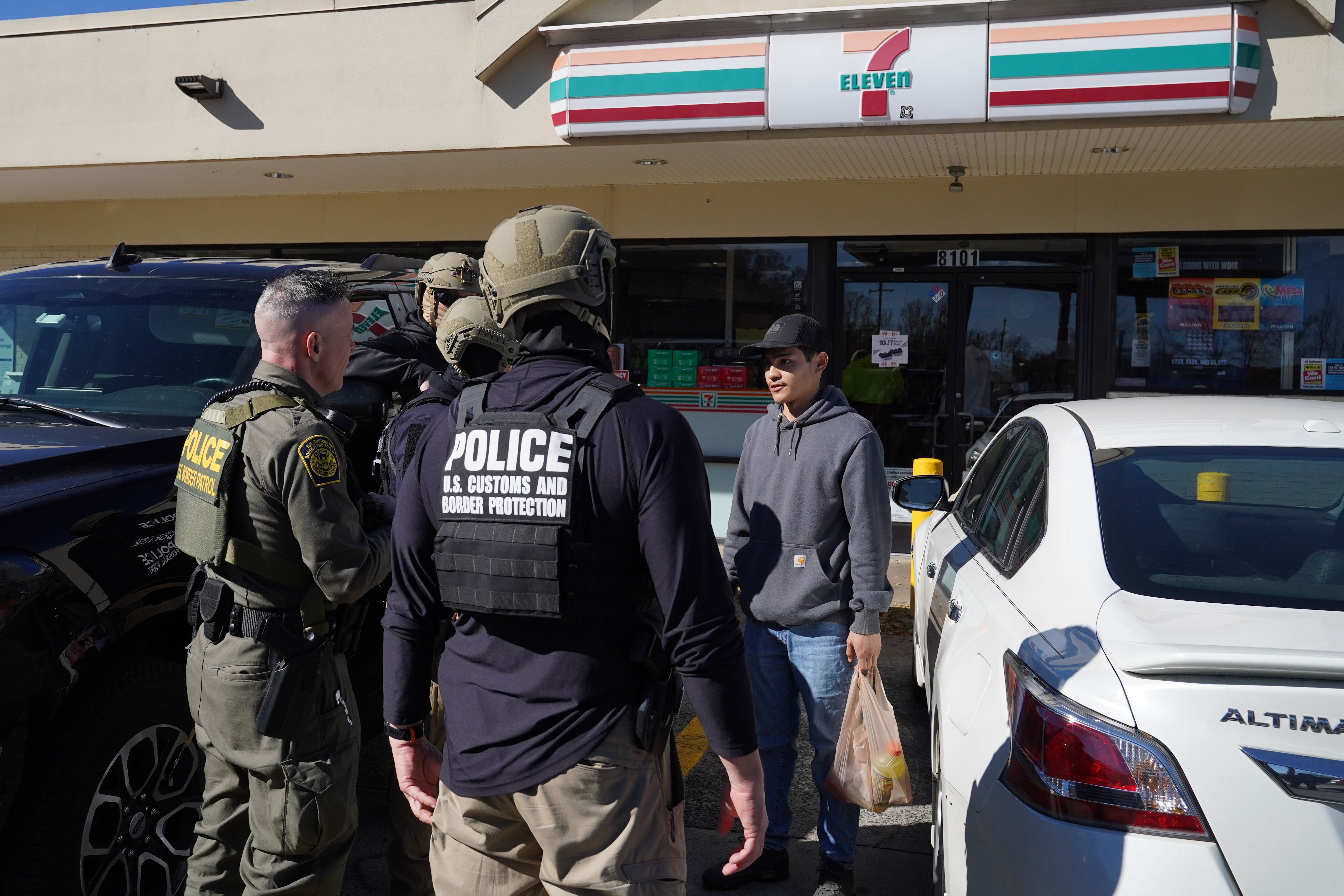 Four U-S Customs and Border Protection officers in tactical gear question a young man in a gray hoodie and black cap holding a bag outside a 7-Eleven.