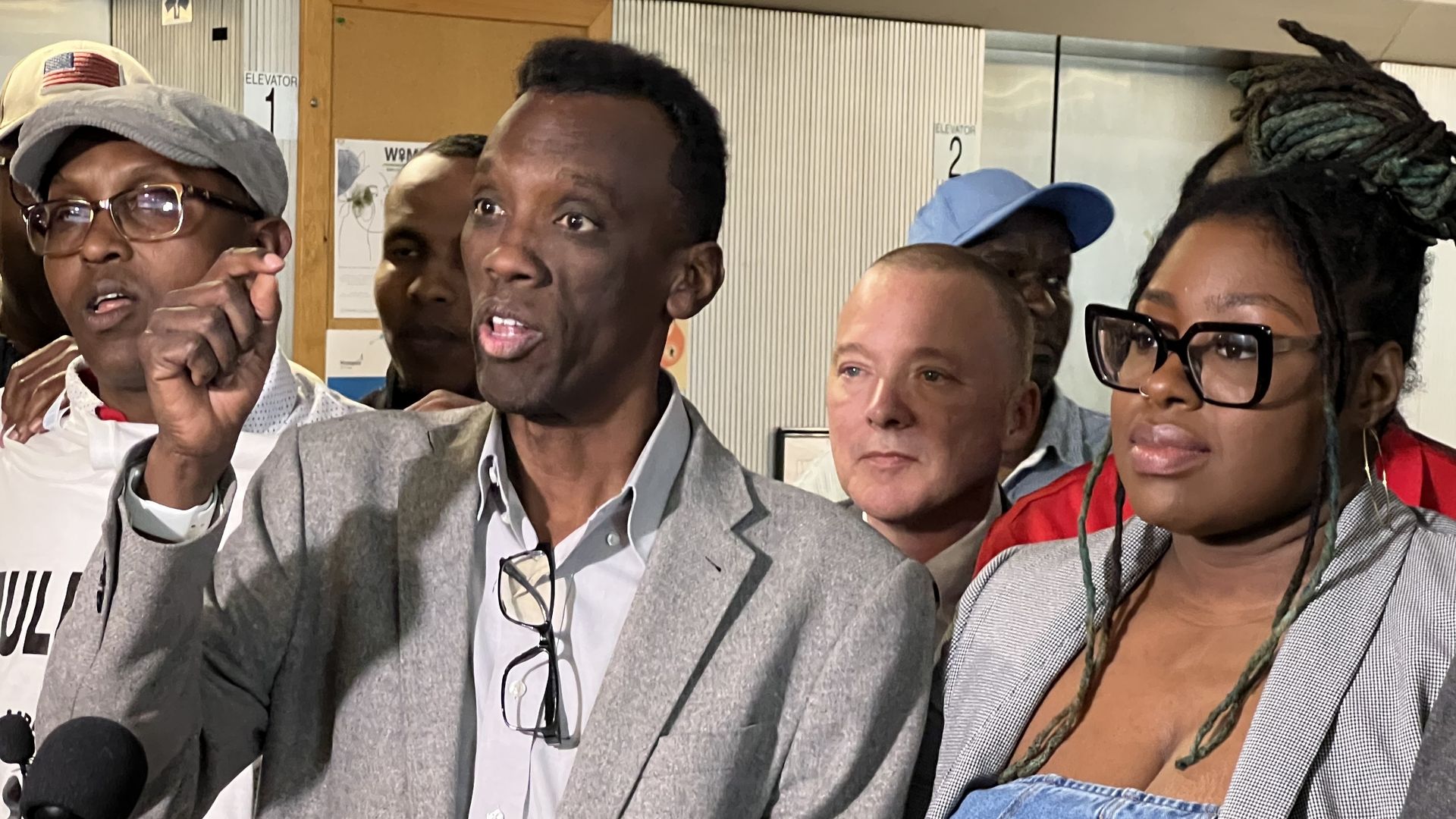 A Black man in a blazer and dress shirt and a Black woman with glasses and a blazer stand at a podium during a press conference. The man on the left is speaking and gesturing with his right hand.