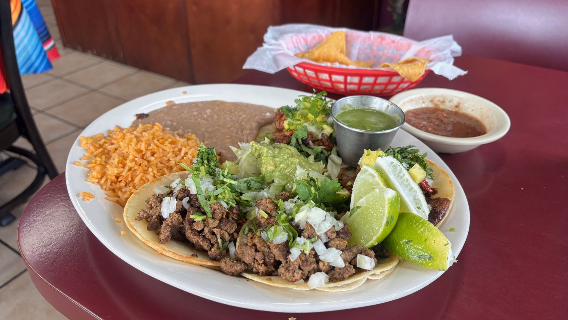 Plate of beef tacos with onions and cilantro, orange rice, beans, guacamole, cilantro, and lime wedges; metal cup of green salsa; red sauce nearby; basket of chips in back.
