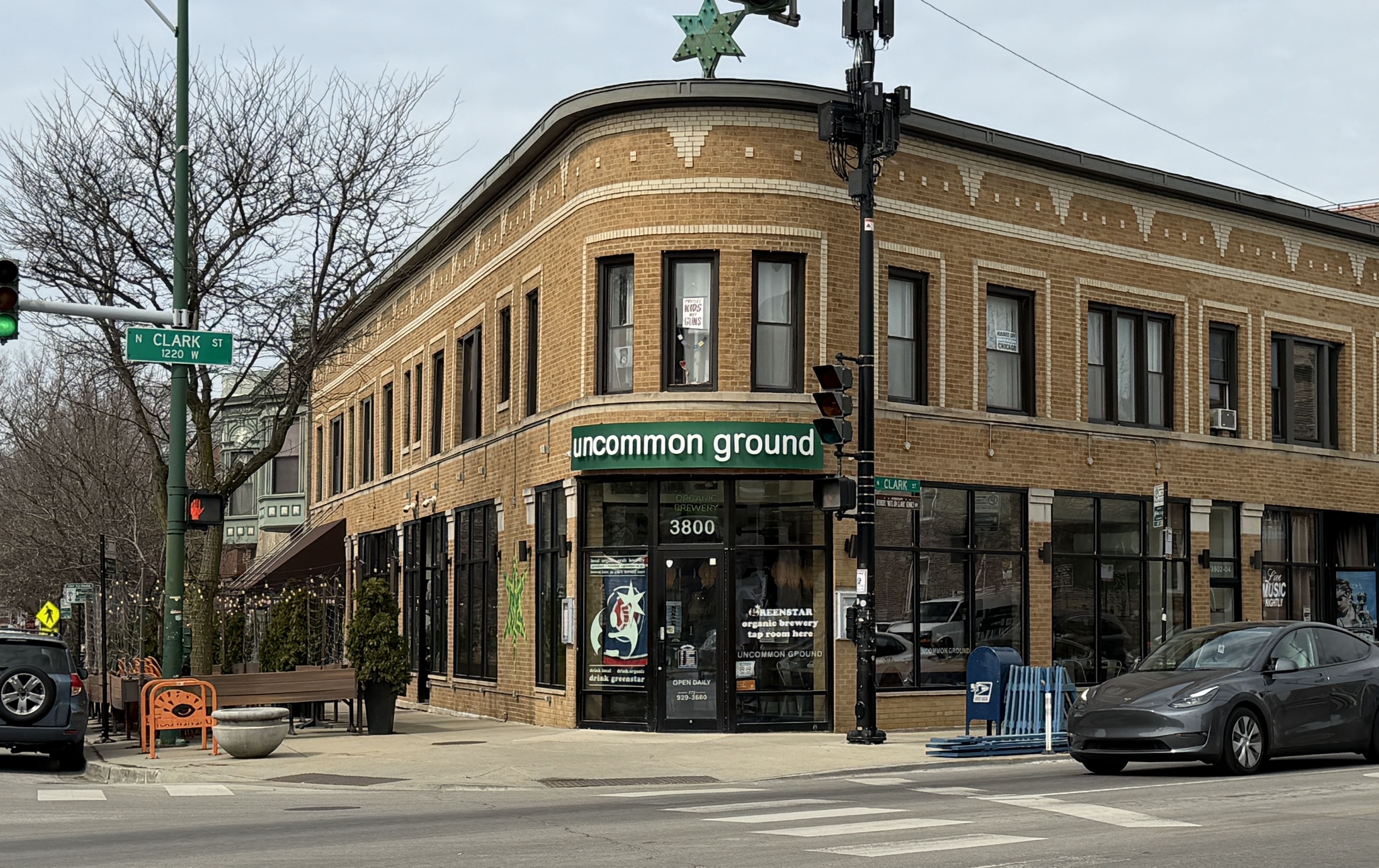 Corner brick building with a green sign reading "uncommon ground" above the storefront; N Clark St sign, crosswalk in front, and a gray Tesla parked at the curb. Leafless trees and outdoor seating line the sidewalk.