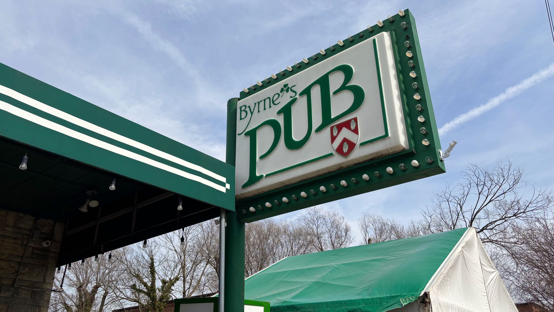 Outdoor street view of a large green-and-white Byrne's Pub sign outlined with bulbs, beside a green awning and white tents; an ATM sign is visible, with leafless trees and a blue sky in the background.