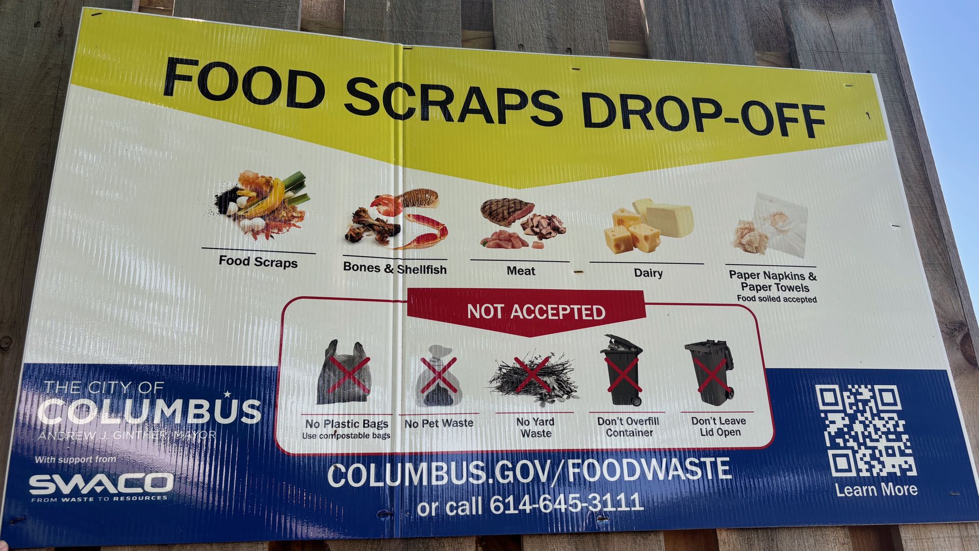 Sign on a wooden fence with a bright yellow banner reading FOOD SCRAPS DROP-OFF; shows food-waste images (meat, cheese, dairy) and a blue sky in the background.