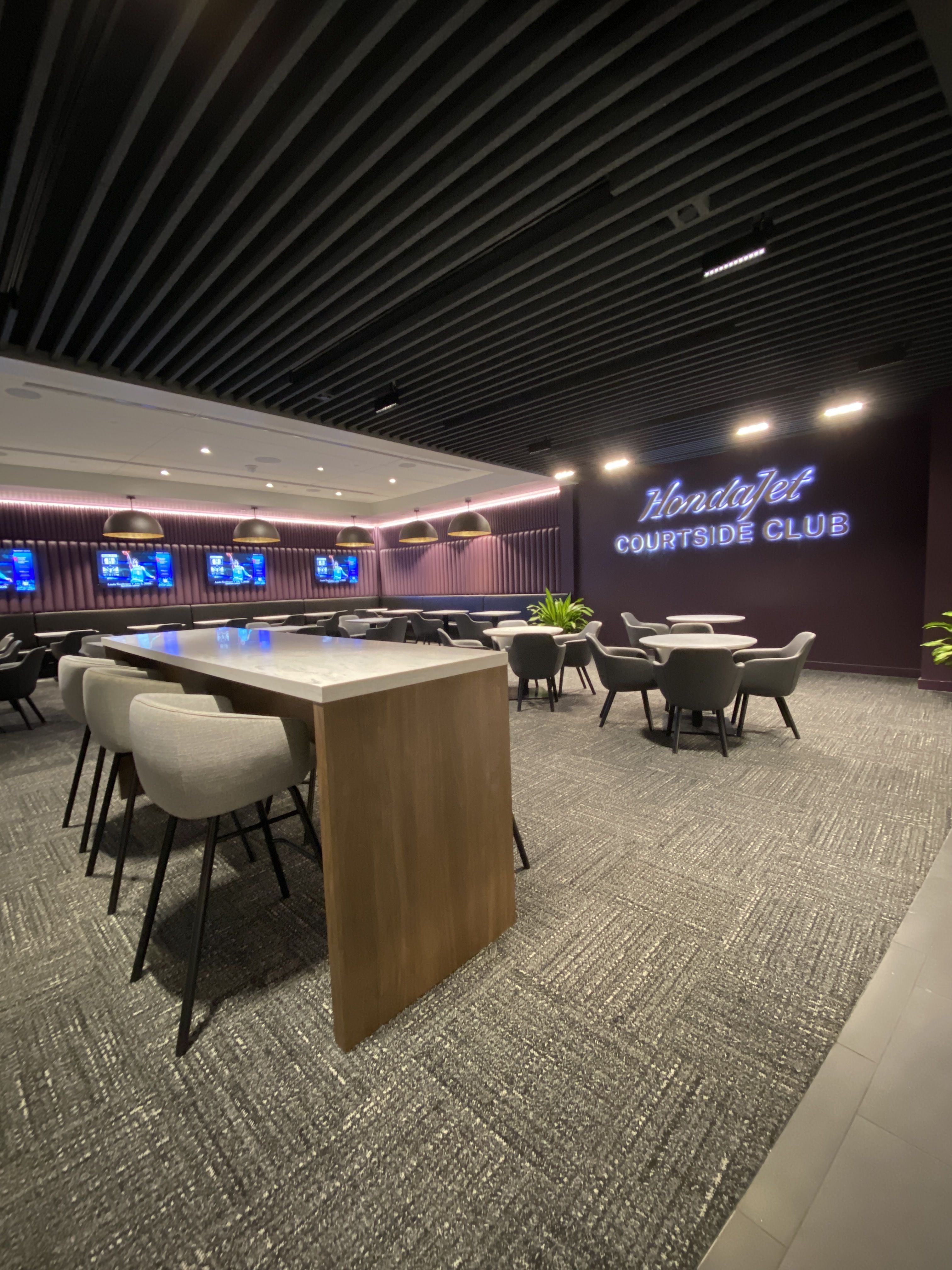 Modern indoor lounge with gray chairs and tables, purple accent walls, multiple TVs, hanging lights, and a glowing sign that reads "HondaJet Courtside Club" on a dark wall.