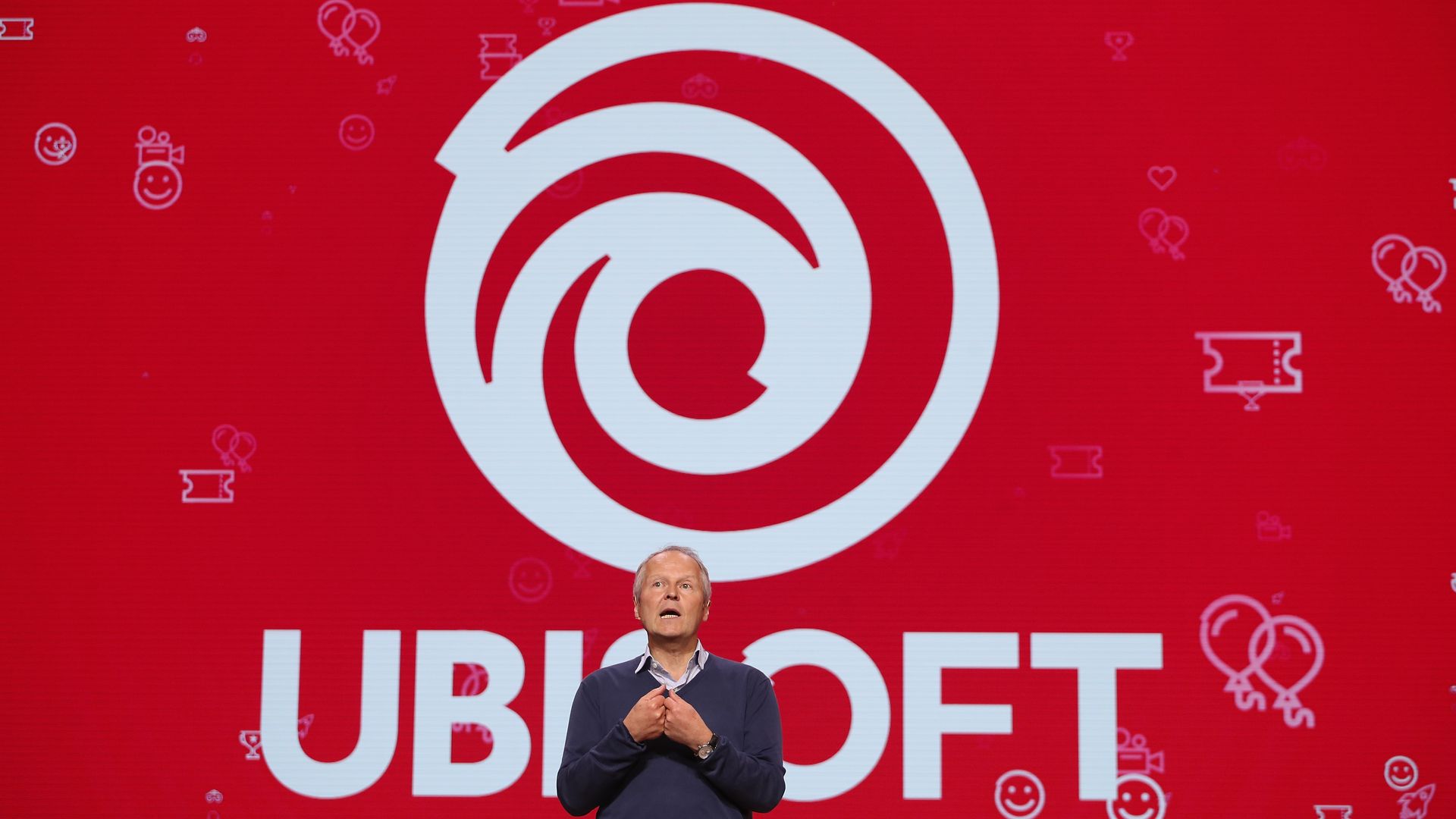 Photo of a man in pants, sweater, and a collard shirt standing in front of a big version of the Ubisoft logo