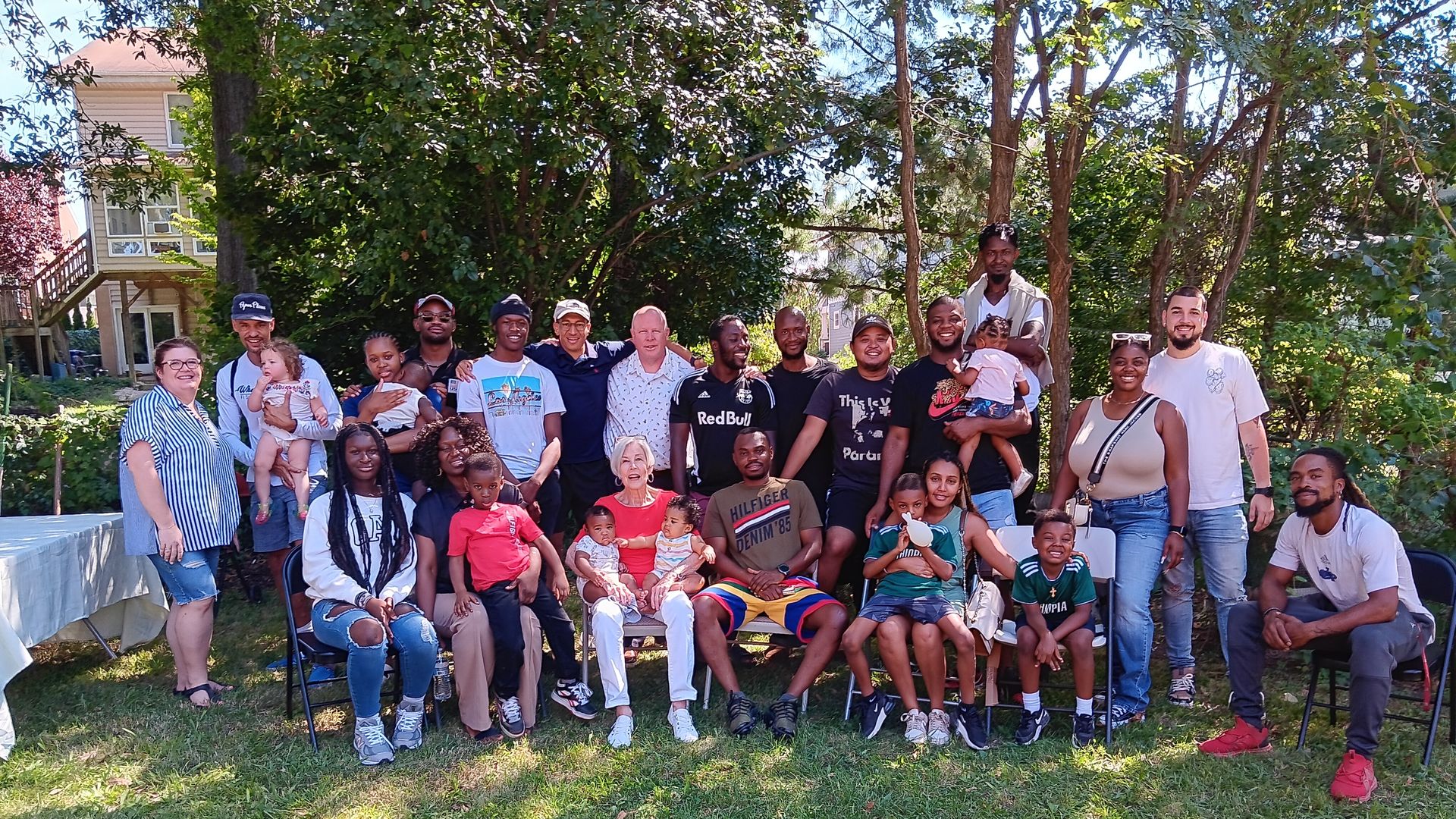 A large group of adults and children pose for a group photo in a sunny backyard, with trees and a house behind them; some sit, others stand, all smiling.