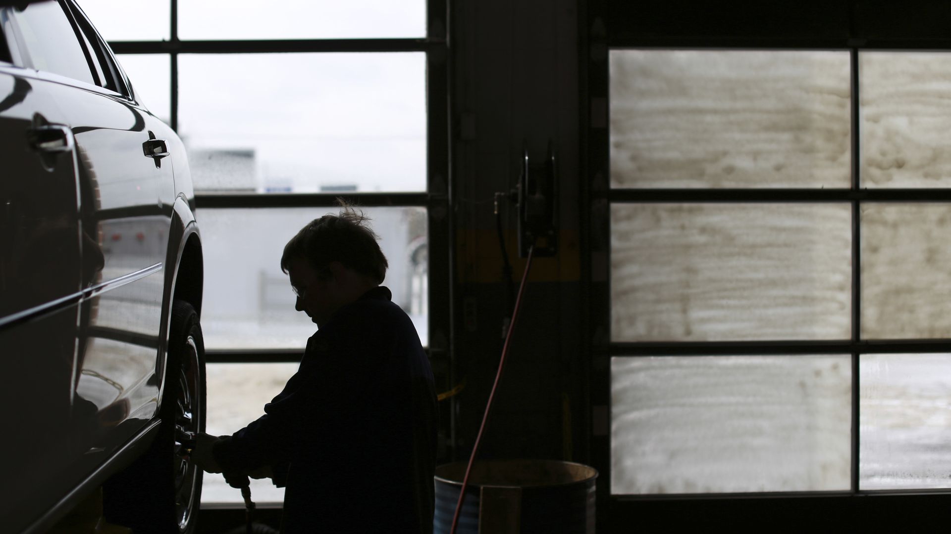 Auto mechanic in shadow working on a raised vehicle with garage door windows in the background.