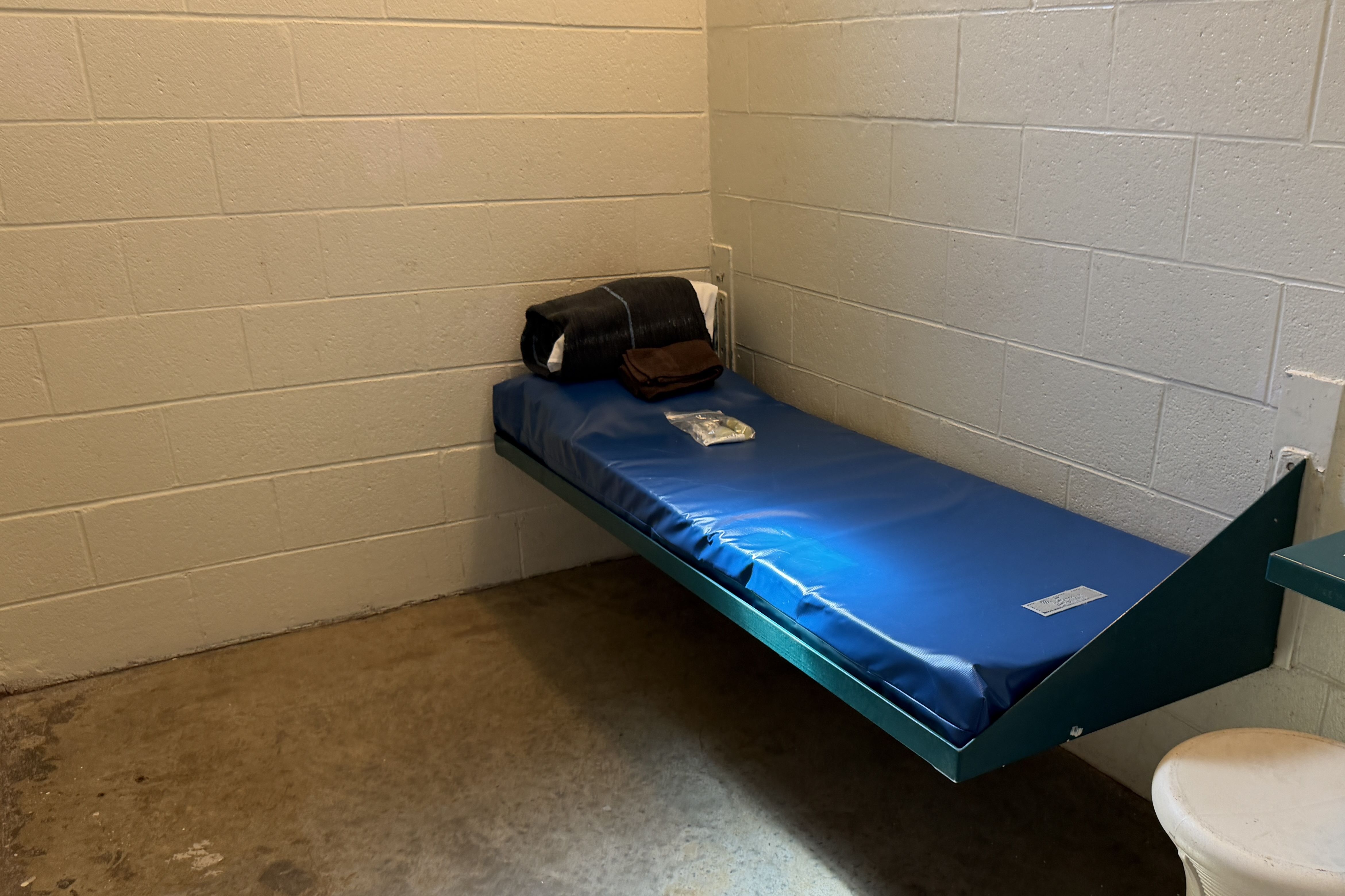 Small room with pale concrete block walls. A blue padded exam table along the right wall holds a folded brown jacket and a dark item, with a small clear plastic bag on the bed.
