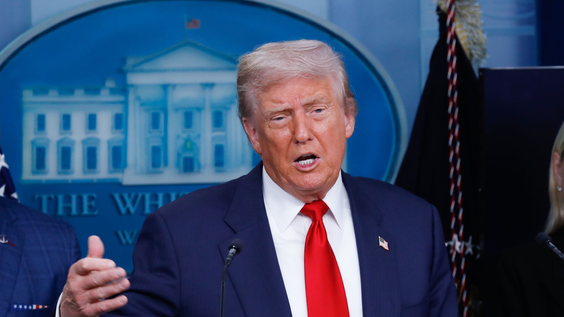President Trump wearing a dark suit and red tie speaking at a podium with the White House emblem in the background.