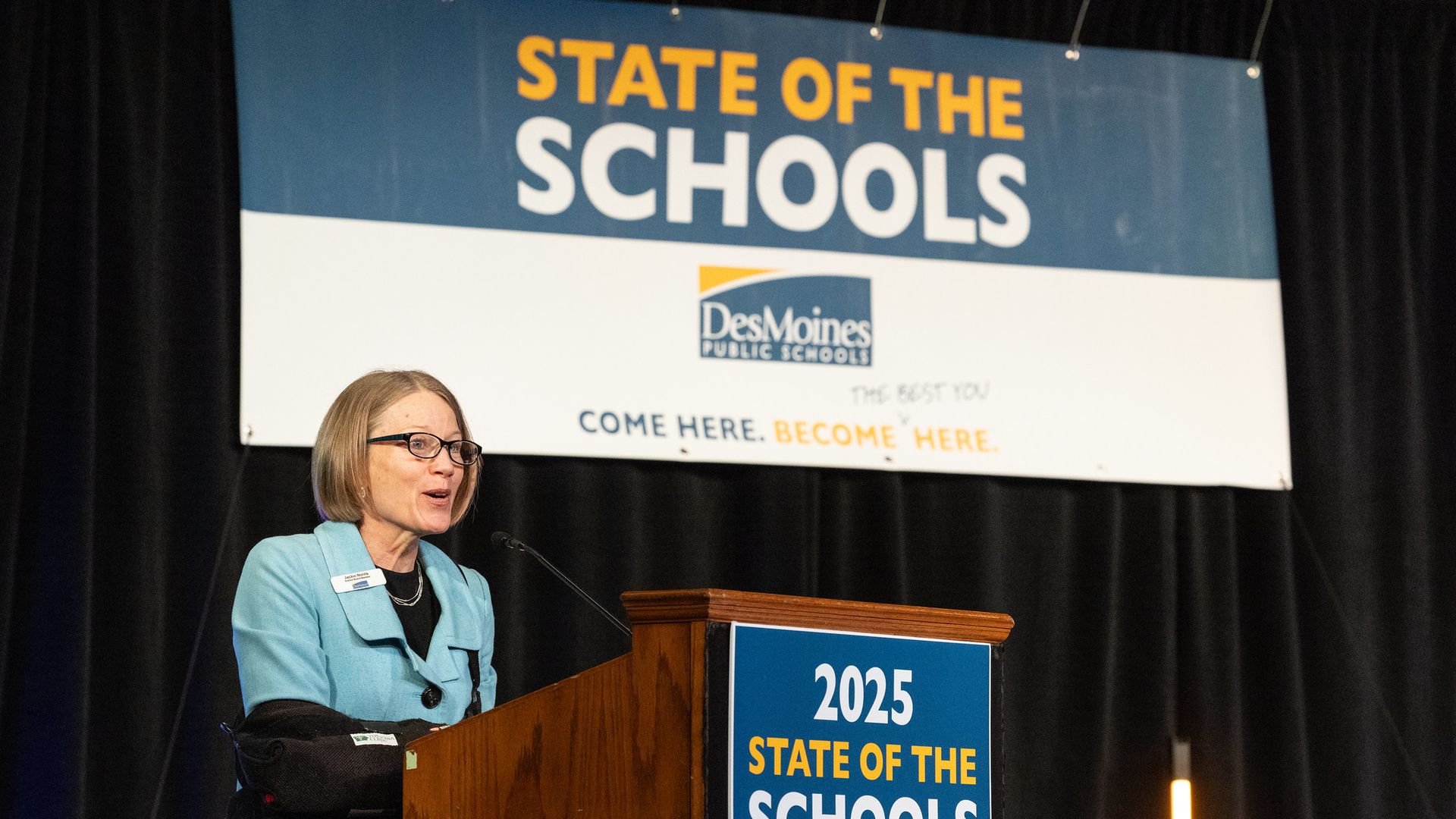 Jackie Norris speaking at a wooden podium with a blue sign reading "2025 State of the Schools" and a banner behind showing "Des Moines Public Schools".