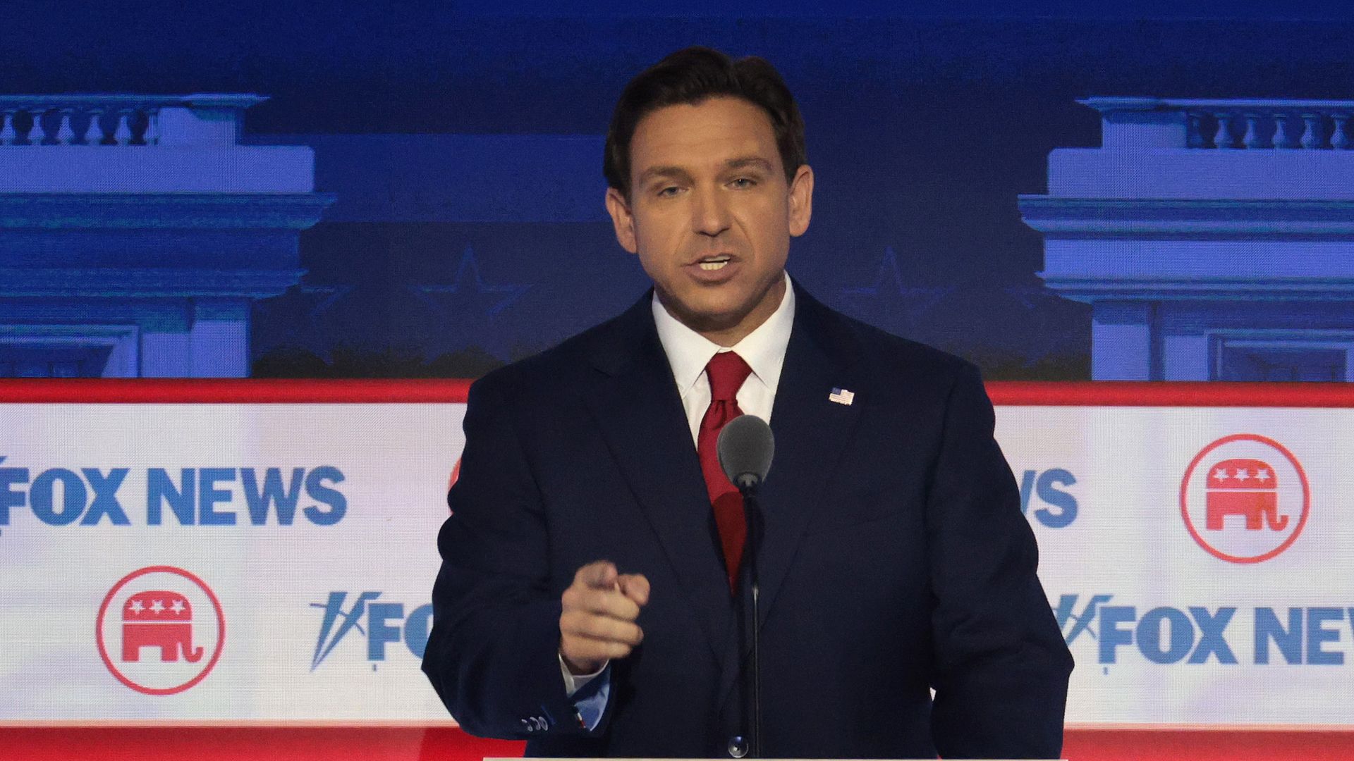 DeSantis in a blue suit and red tie in front of banners that say Fox News stands at a lectern speaking