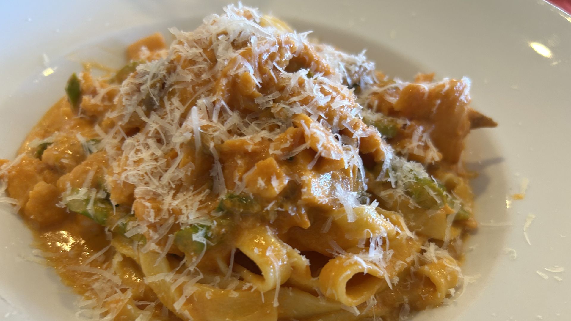 Close-up of a pasta dish in a white bowl: orange creamy sauce with vegetables and pasta, generously topped with shredded white cheese.