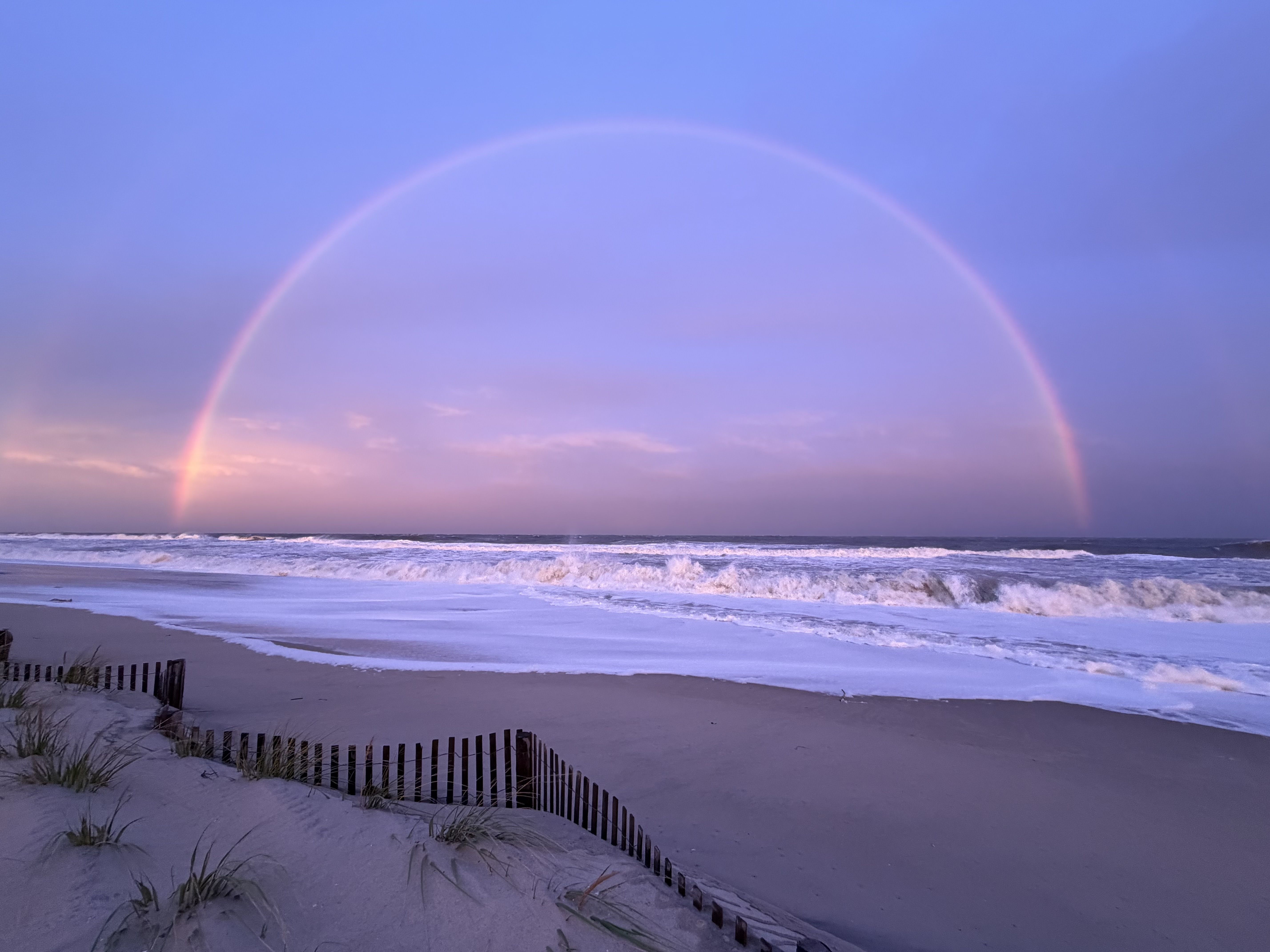 A full rainbow stretches across a purple-blue sky above crashing waves and a sandy beach in Harvey Cedars, N.J.