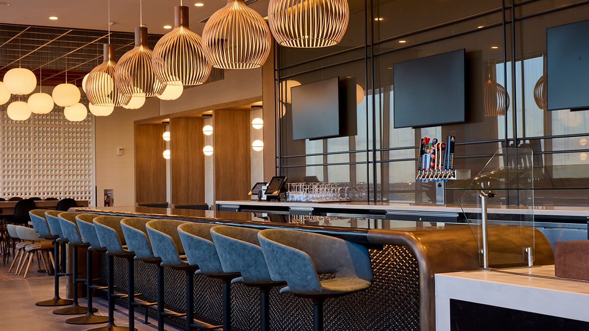 Modern bar interior with blue cushioned stools, pendant lights, multiple flat-screen TVs, and beer taps, featuring a warm color palette and clean, sleek design.