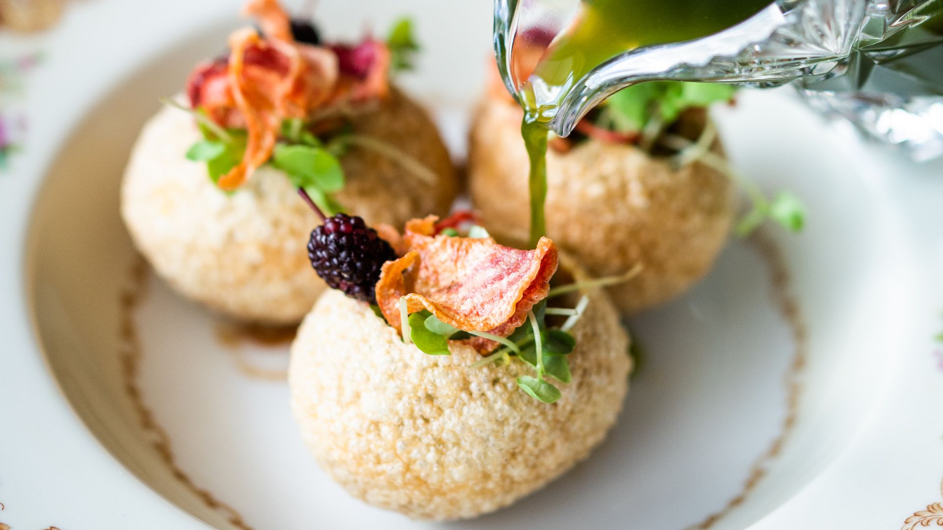 A close-up photo of a green liquid pouring from a crystal pitcher into one of three puffed, garnished spheres.