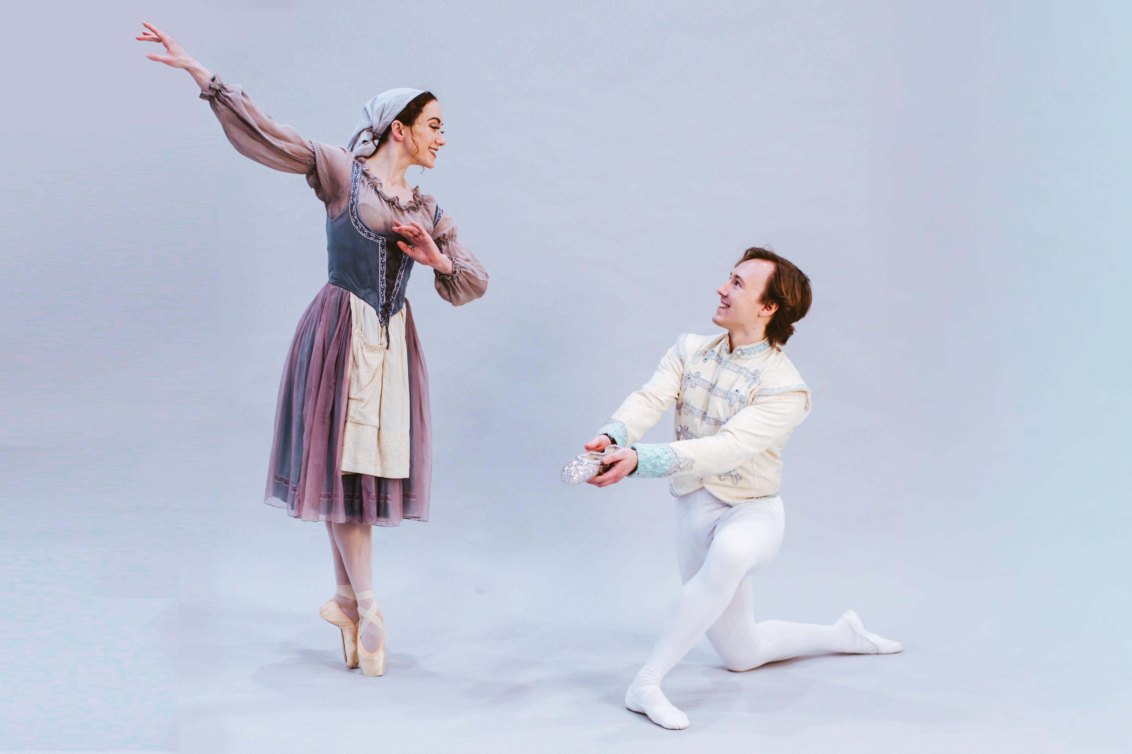 Two ballet dancers in period costumes perform a duet: a woman on pointe with arms extended in a dusty pink and blue gown, and a kneeling man in white offering a glittery prop against a pale blue backdrop.