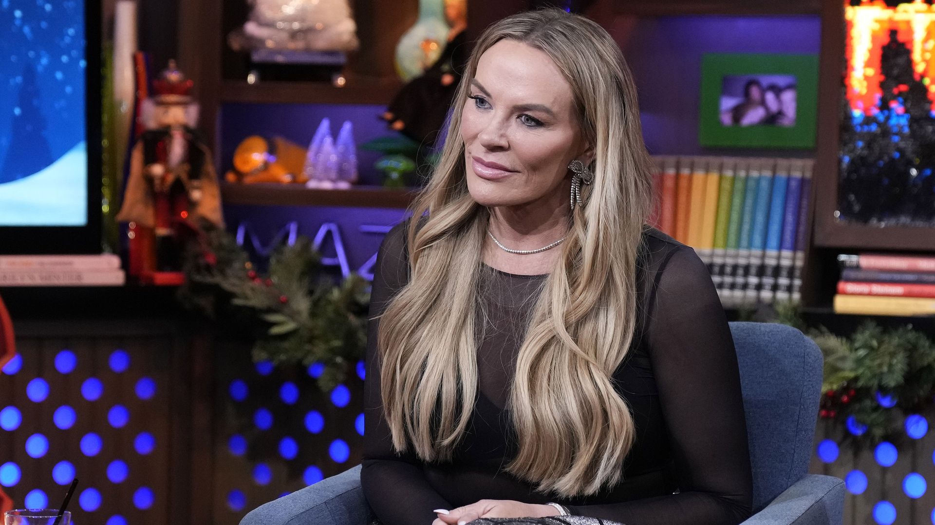 Woman with long blonde hair in a black sheer top and silver skirt sitting in a blue armchair during a festive indoor interview setting with colorful books and holiday decorations.