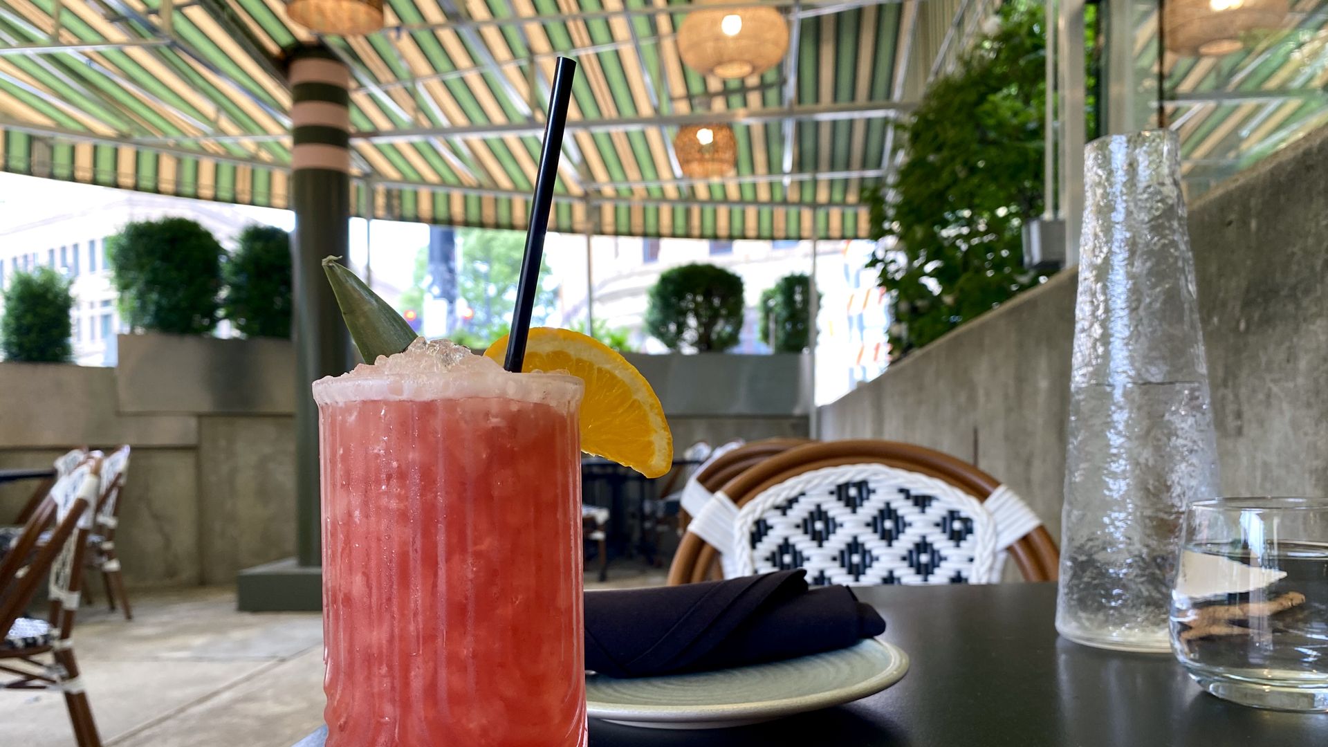 Pink cocktail with crushed ice, a black straw, orange wheel, and pineapple leaf on a dark table. Outdoor cafe setting with a green striped awning, woven lanterns, and wicker chairs.