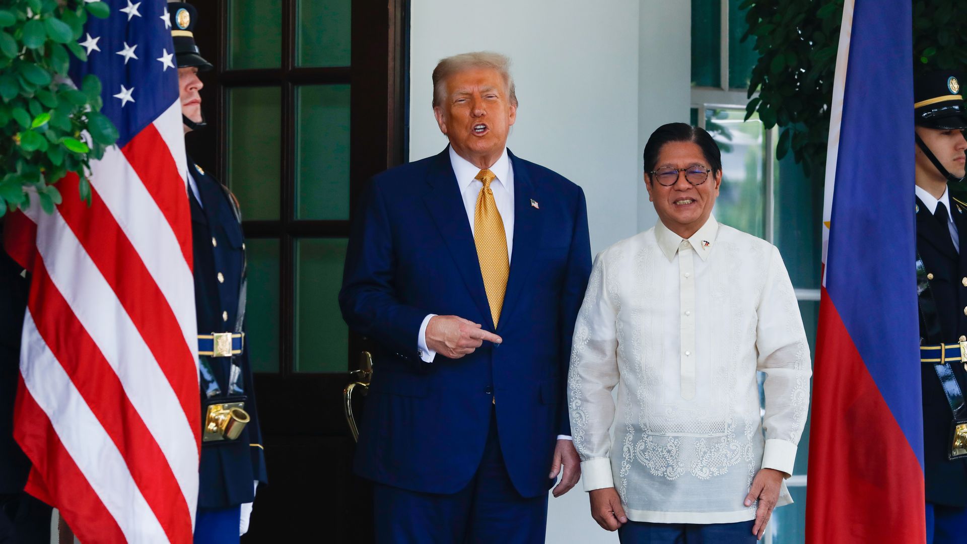 Two men stand side by side between American and Philippine flags, flanked by uniformed guards. The man on left wears navy suit, yellow tie, the other wears white barong tagalog shirt.