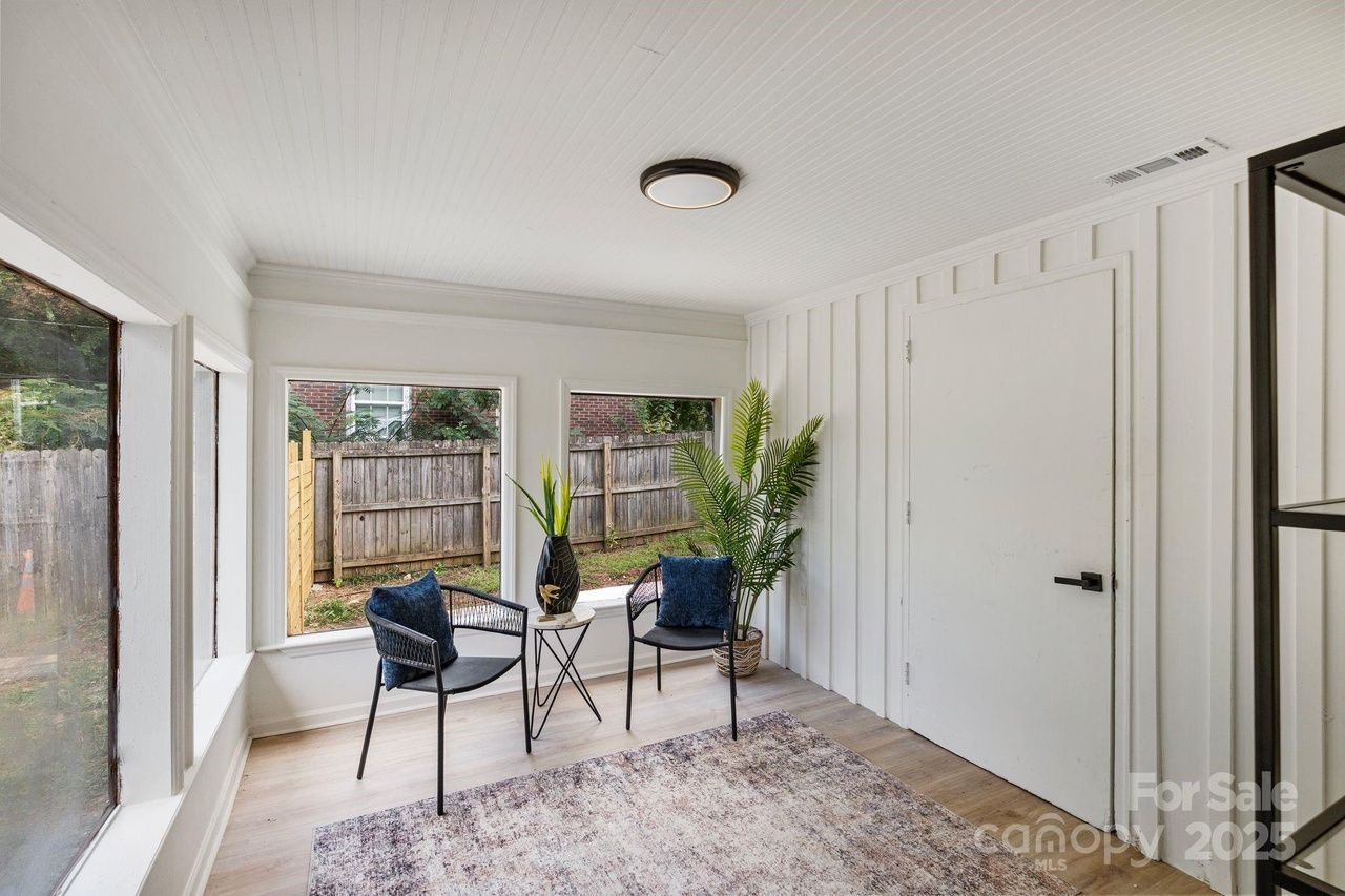 Small sunroom with white walls and ceiling, two black chairs with blue cushions, a small round white table, potted green plant, large windows showing wooden fence and backyard.