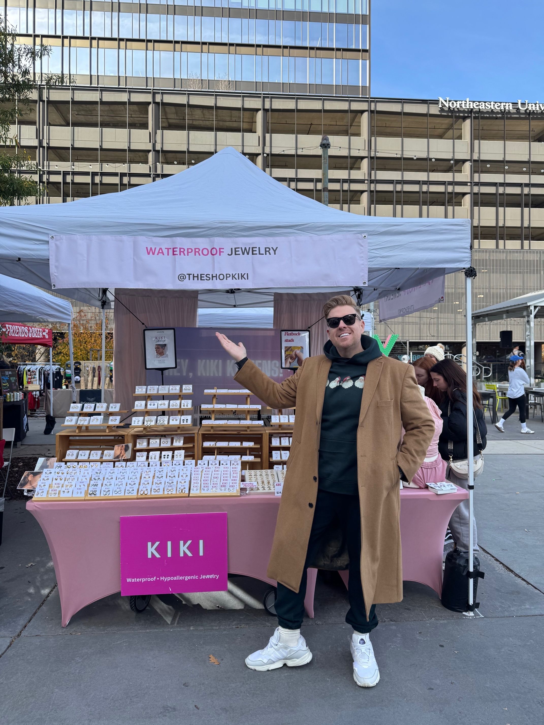 Man in brown coat and sunglasses smiles and gestures to a jewelry stall with a white canopy. The stall displays waterproof jewelry and has a pink sign reading "KIKI Waterproof Hypoallergenic Jewelry."