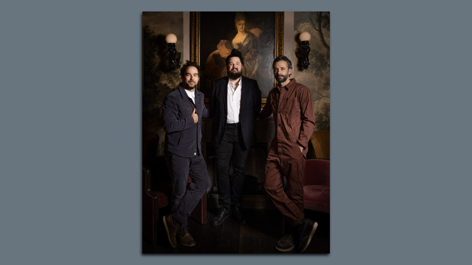 Three men pose indoors against a wall with vintage wallpaper and a large framed painting of a woman. The man left wears dark blue, center wears black suit, right wears brown outfit.