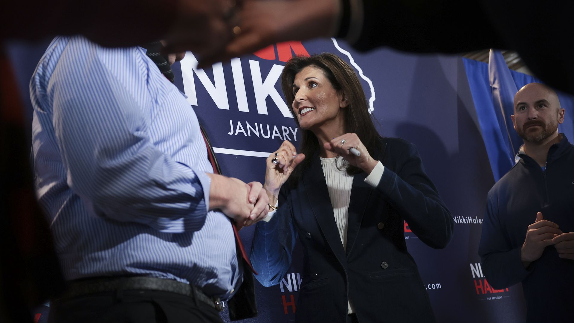 Nikki Haley, in a blue blazer and white blouse, talks with a supporter on a podium and in front of a banner.