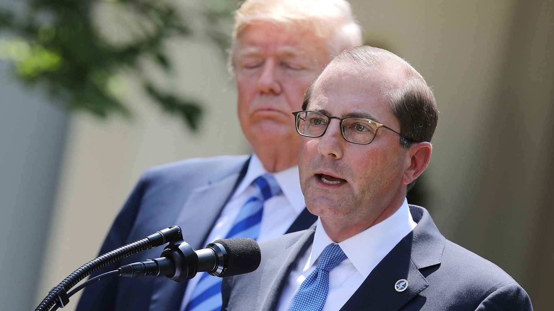 Azar stands behind a microphone while Trump stands behind him