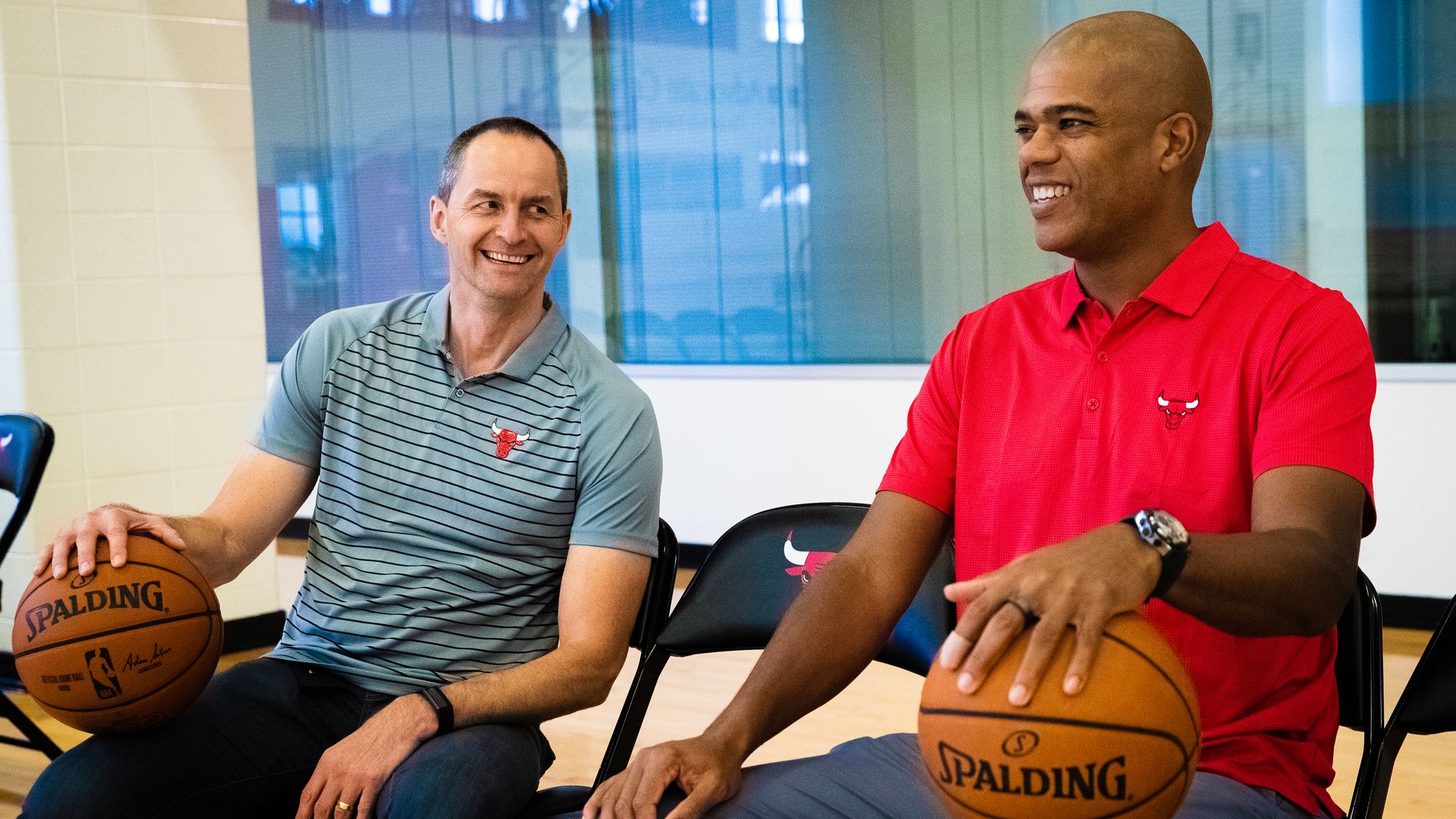 Two men sit on folding chairs in a bright gym, smiling while each holds a Spalding basketball. Left wears a teal striped polo; right wears a red polo with a Bulls logo.