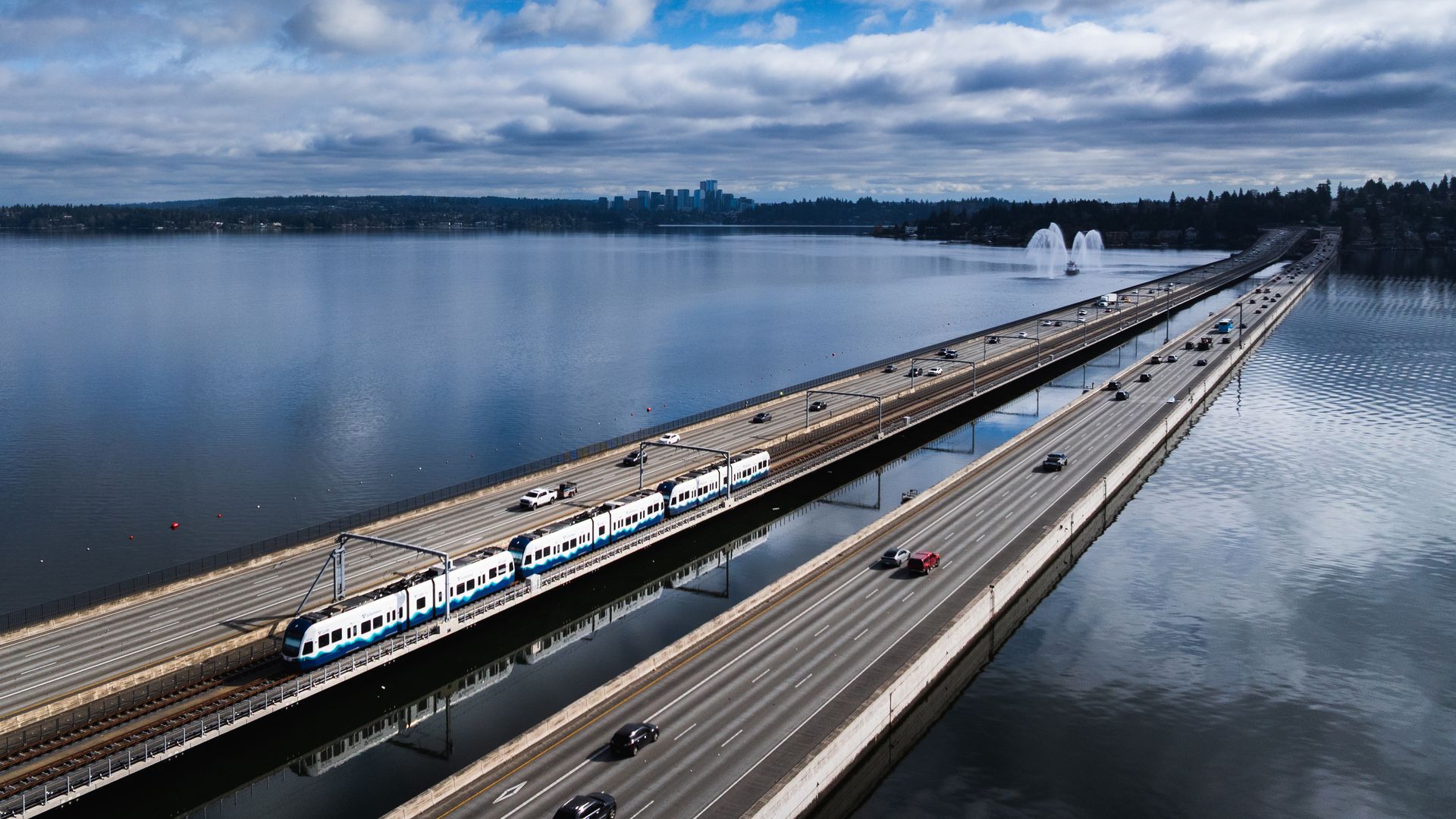Wide view of a bridge over calm water with a blue and white commuter train, cars in both directions, a fountain in the water, and a distant city skyline under cloudy skies.