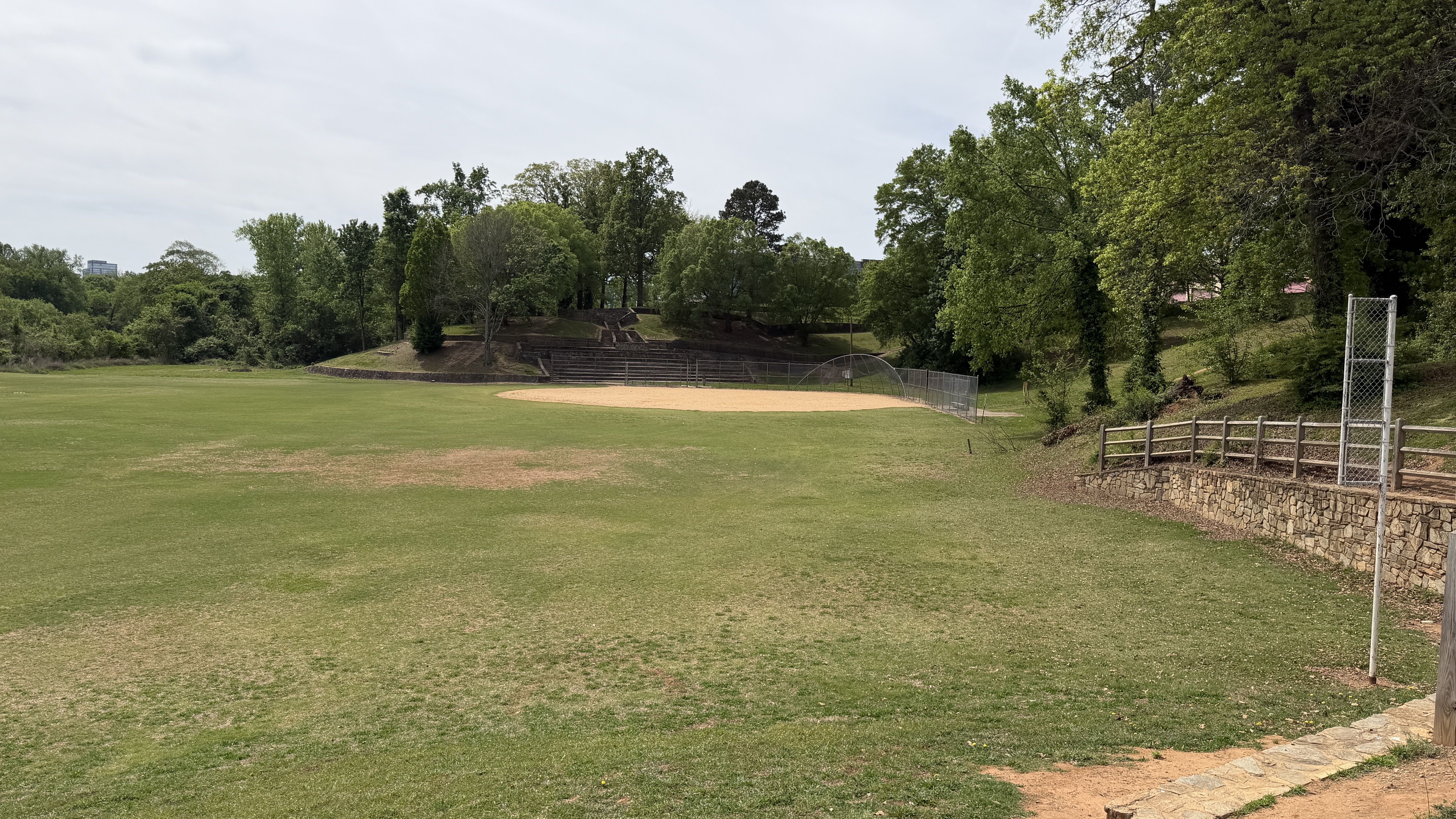 View from a stone overlook of a baseball diamond in a park: dirt infield, green outfield, chain-link fence, surrounding trees, and a blue sky with wispy clouds.