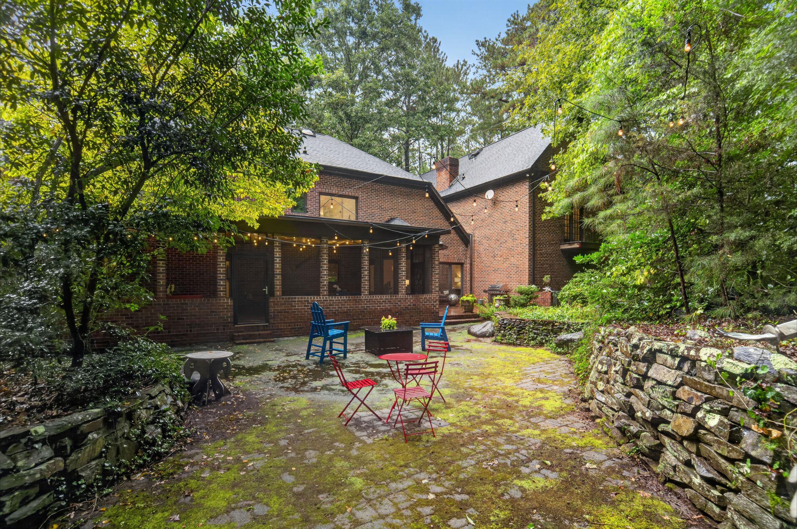 Brick house with a slate roof sits in a leafy backyard. String lights crisscross above a mossy stone patio, where red and blue metal chairs are arranged among trees and rock walls.