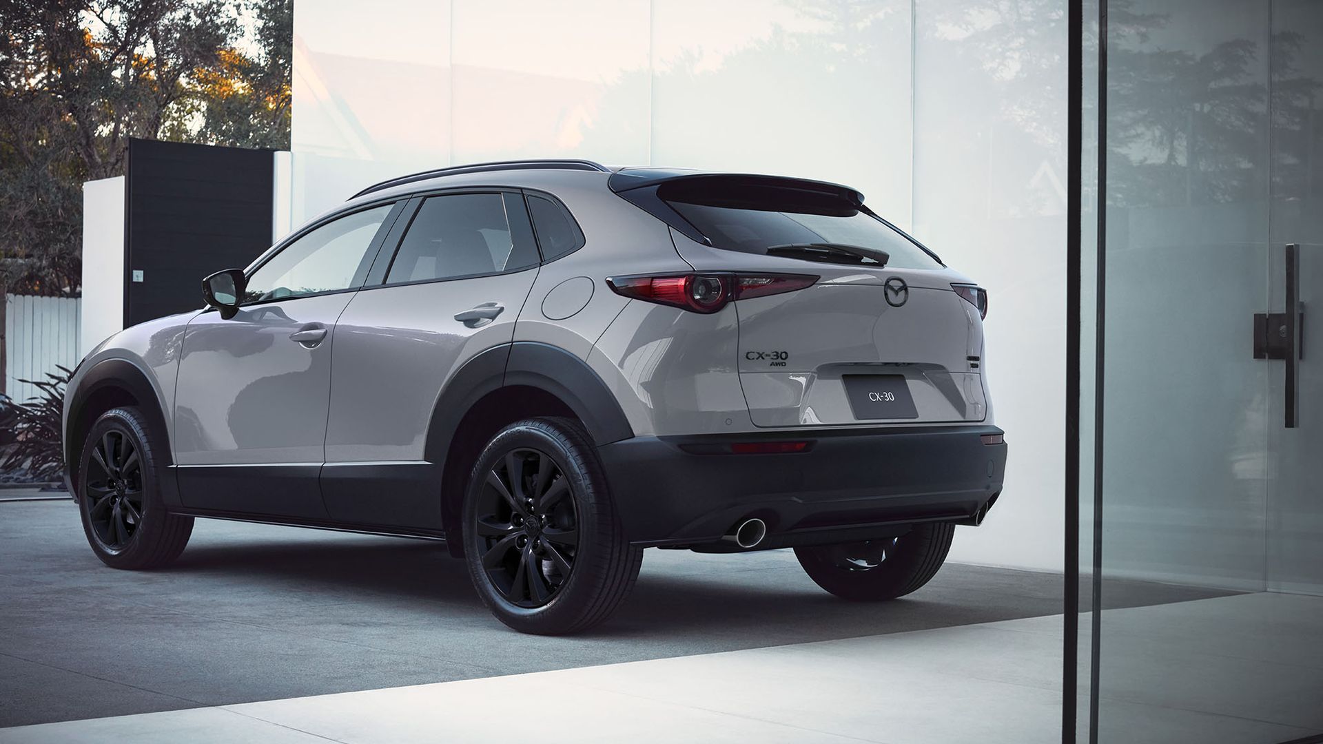 Rear-side view of a silver Mazda CX-30 AWD SUV parked in front of a modern glass building; visible taillights, rear badge, and reflections on the glass.