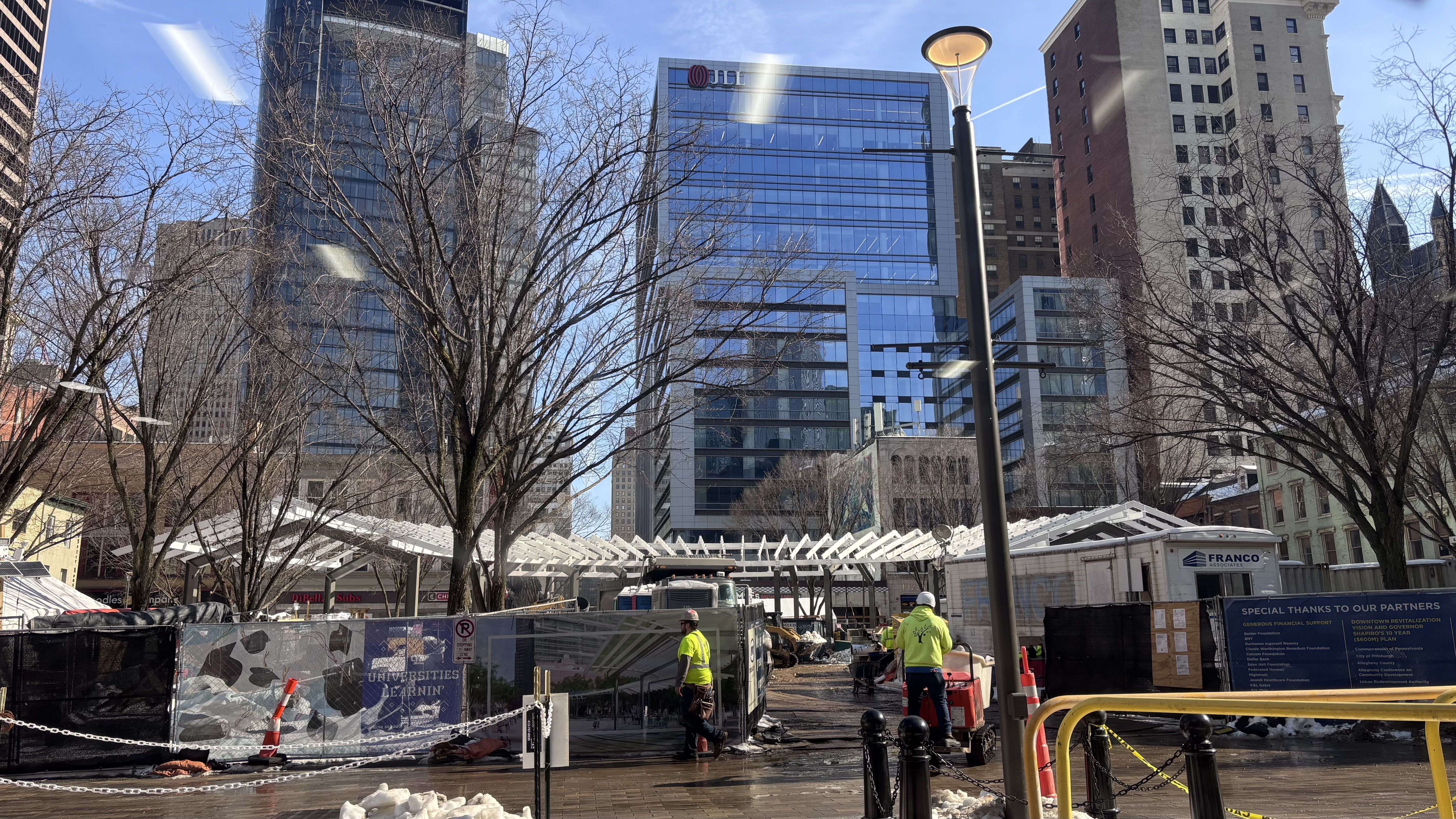 Busy city construction site with workers in neon yellow jackets, leafless trees, tall glass and brick buildings in the background under a clear blue sky.