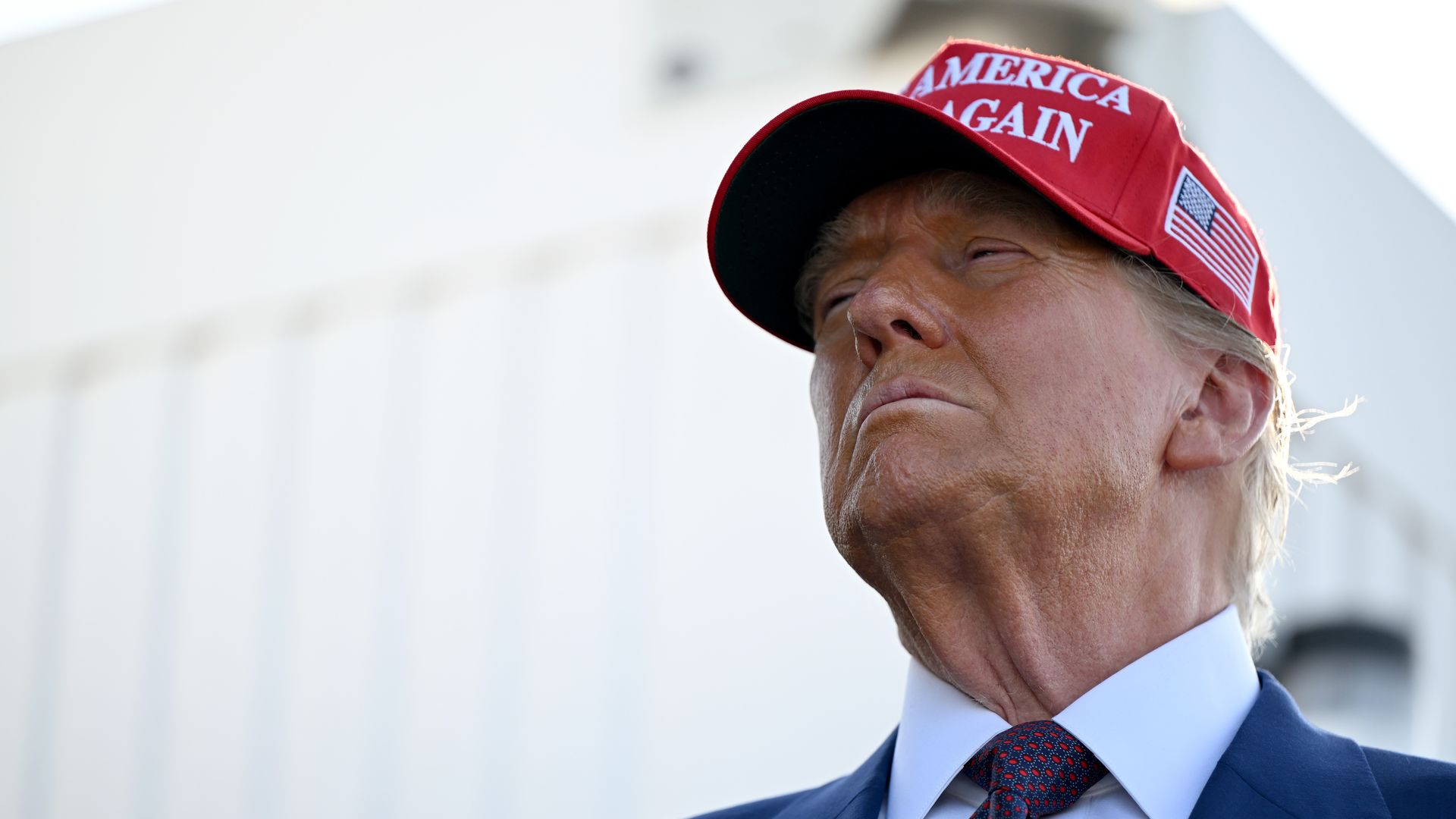 A photo of Trump taken from below his face. He wears a red MAGA hat. 