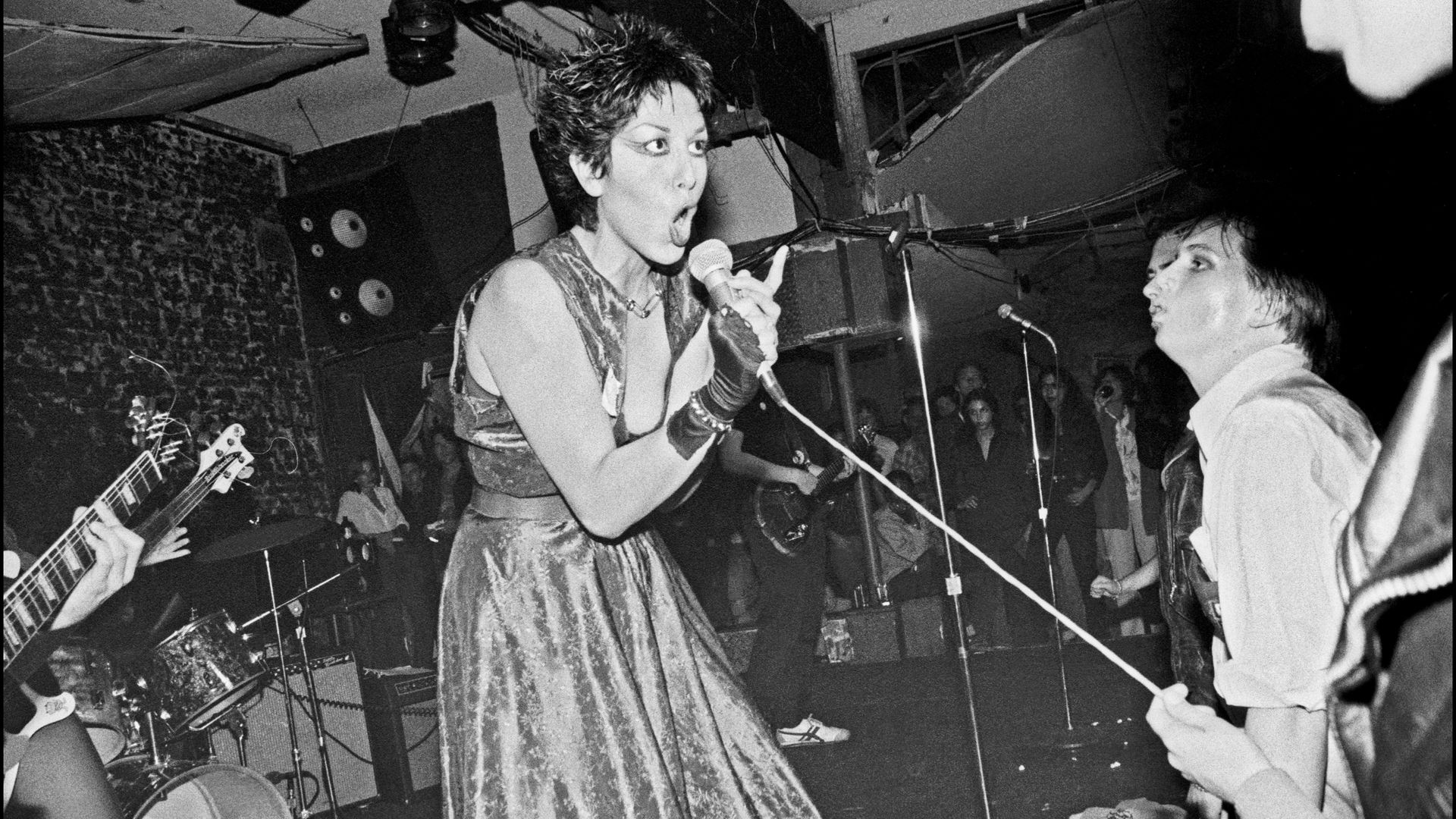 Photo of Alice Bag singing into a microphone while a band players behind her and people watch from below the stage
