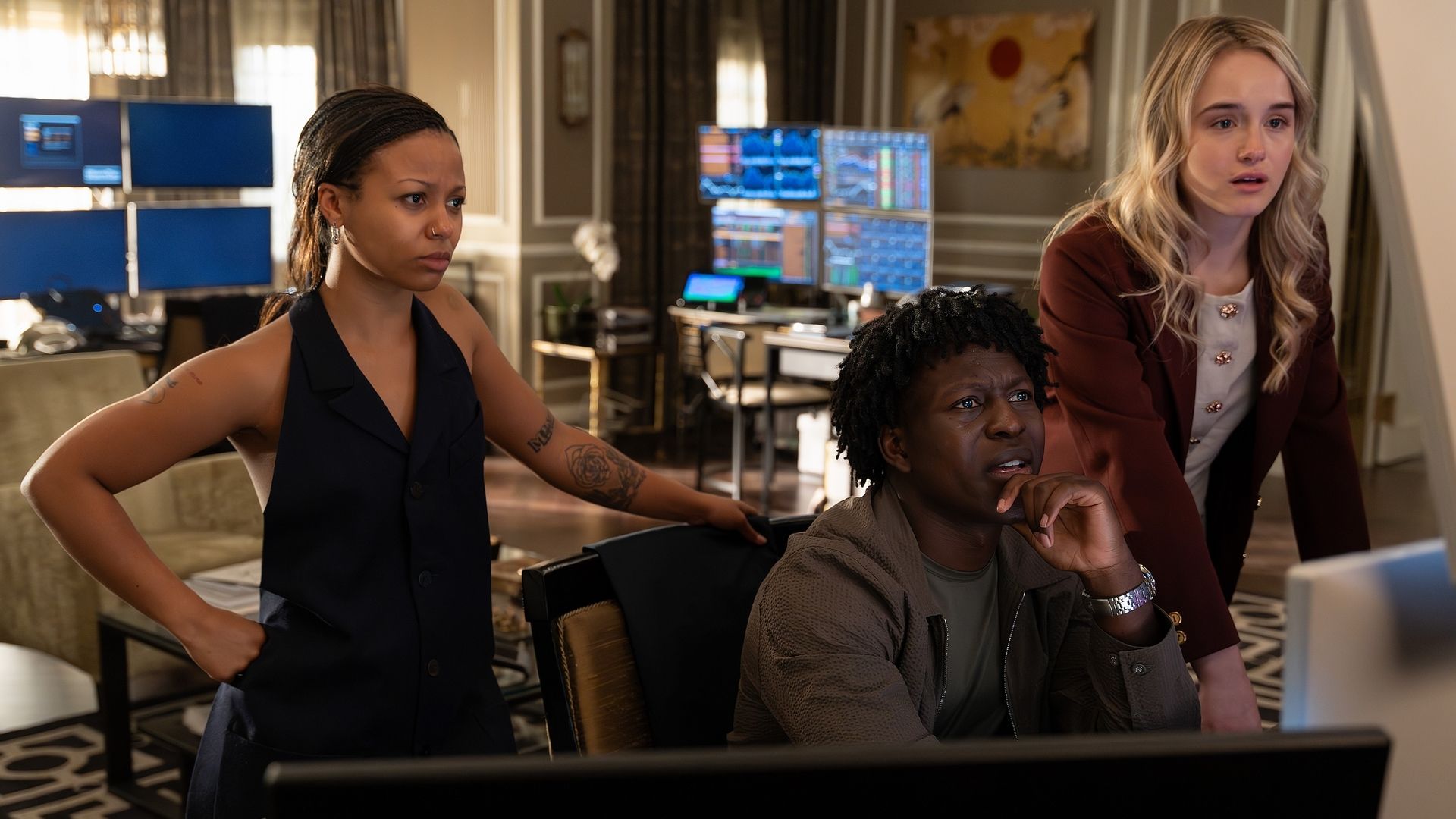 Three people focused on a computer screen in a room with multiple monitors displaying charts and data. A woman in black stands, a man seated with hand on chin, and a blonde woman in maroon leans forward.