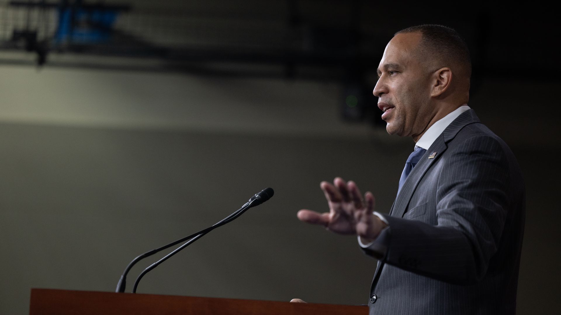 Hakeem Jeffries speaking at a podium in front of a gray wall wearing a gray suit.