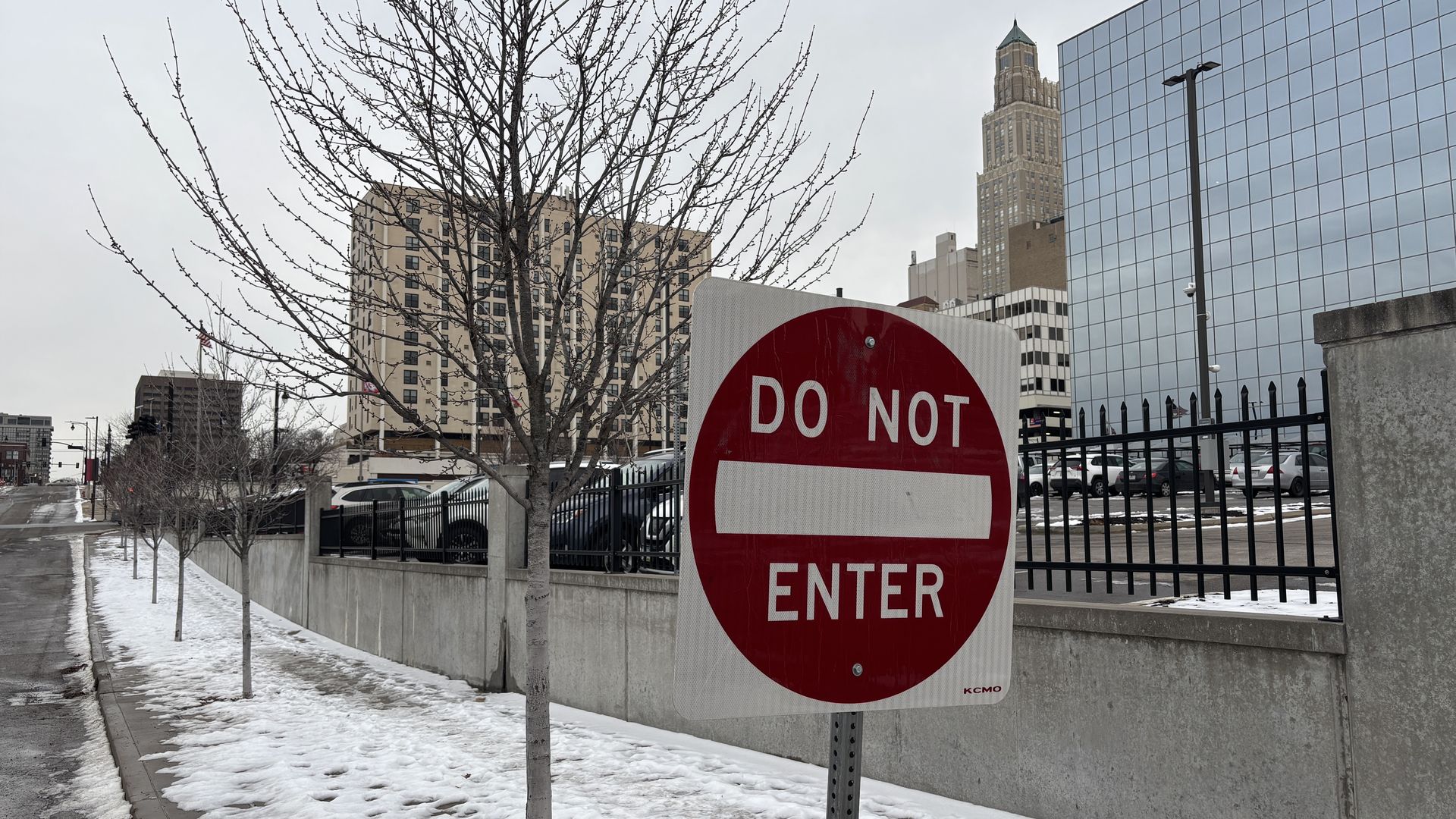Red and white "DO NOT ENTER" sign beside a snowy sidewalk with bare trees, a black fence, parked cars, and tall buildings in the background under a cloudy sky.