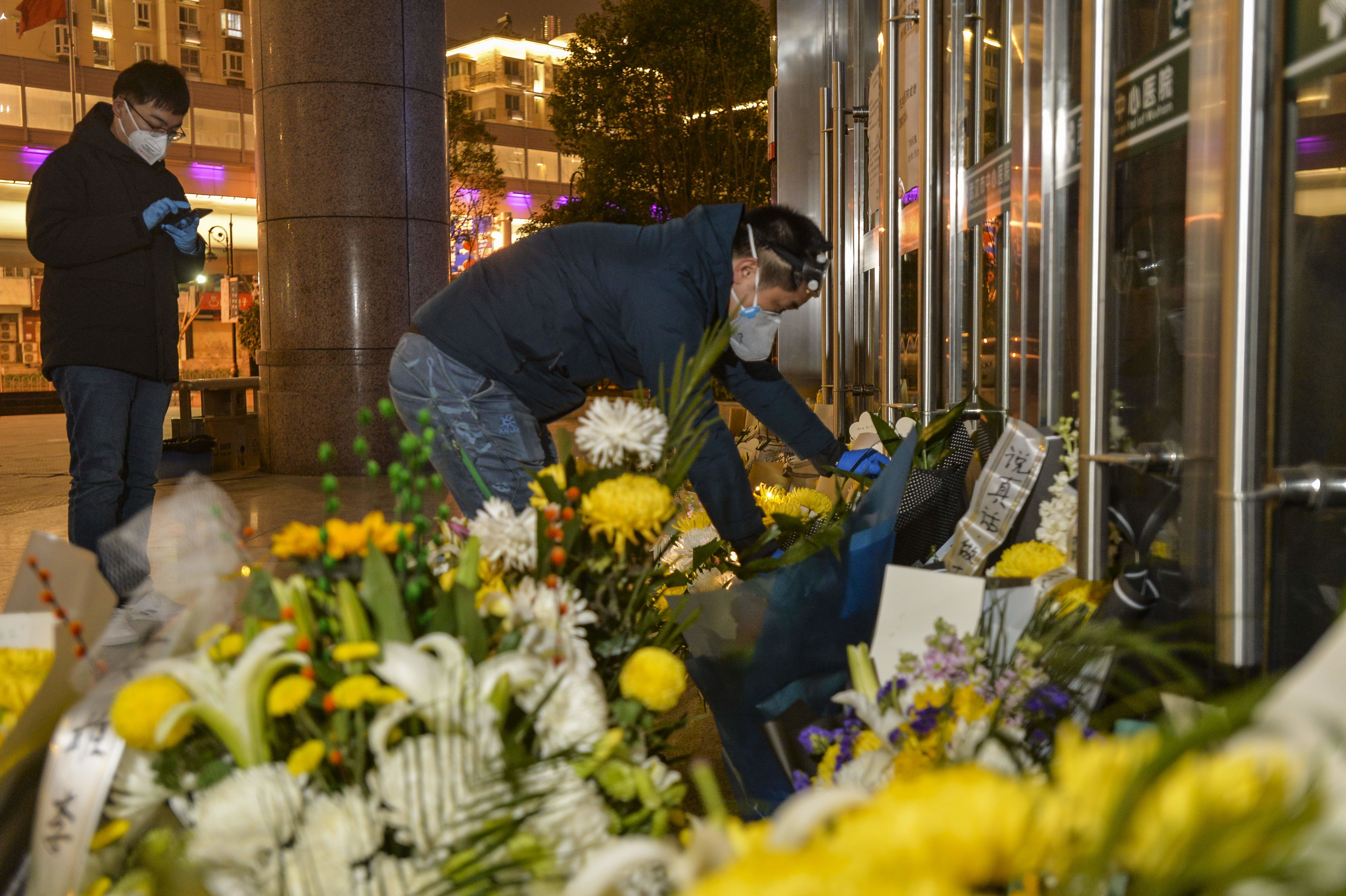 In this image, a person wearing a protective face mask places flowers against the front glass doors of a building