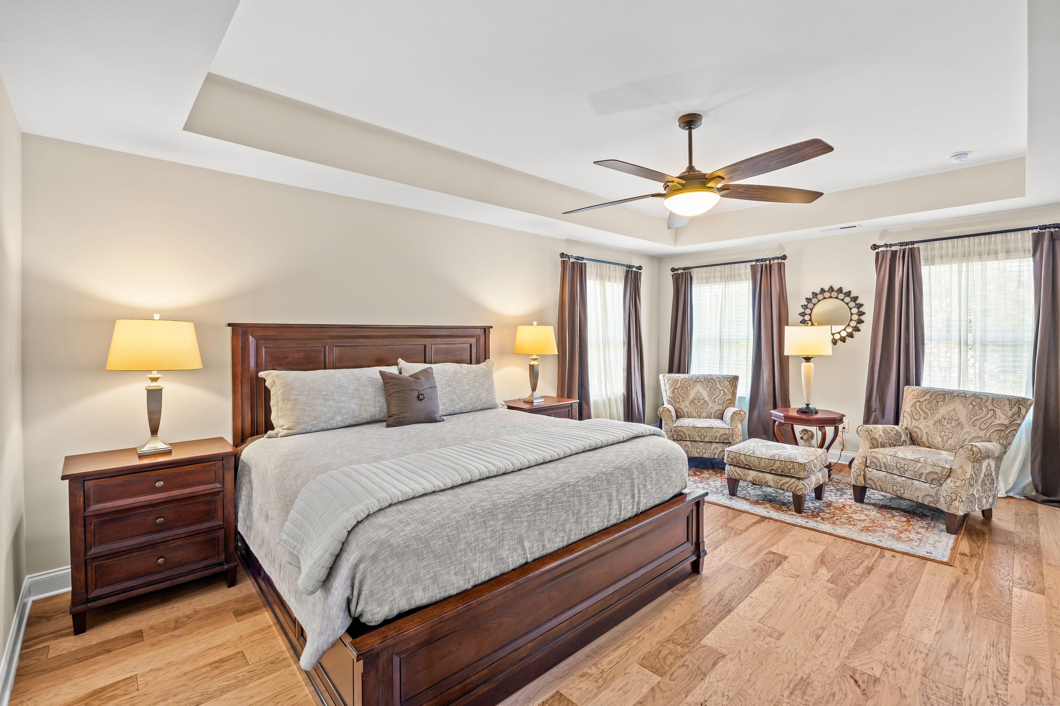 Spacious bedroom with dark wood bed, matching nightstands, beige walls, and a ceiling fan. A seating area with two patterned chairs, an ottoman, rug, and large windows with brown curtains.