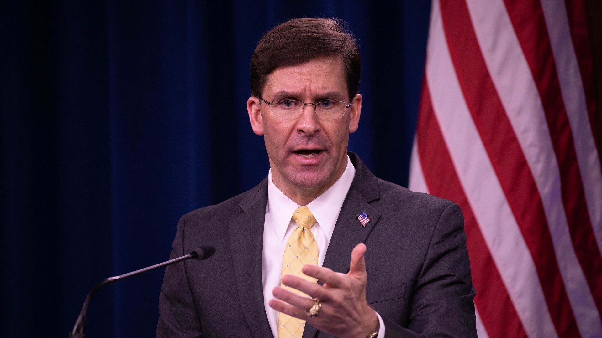 In this image, Mark Esper stands in front of an American flag