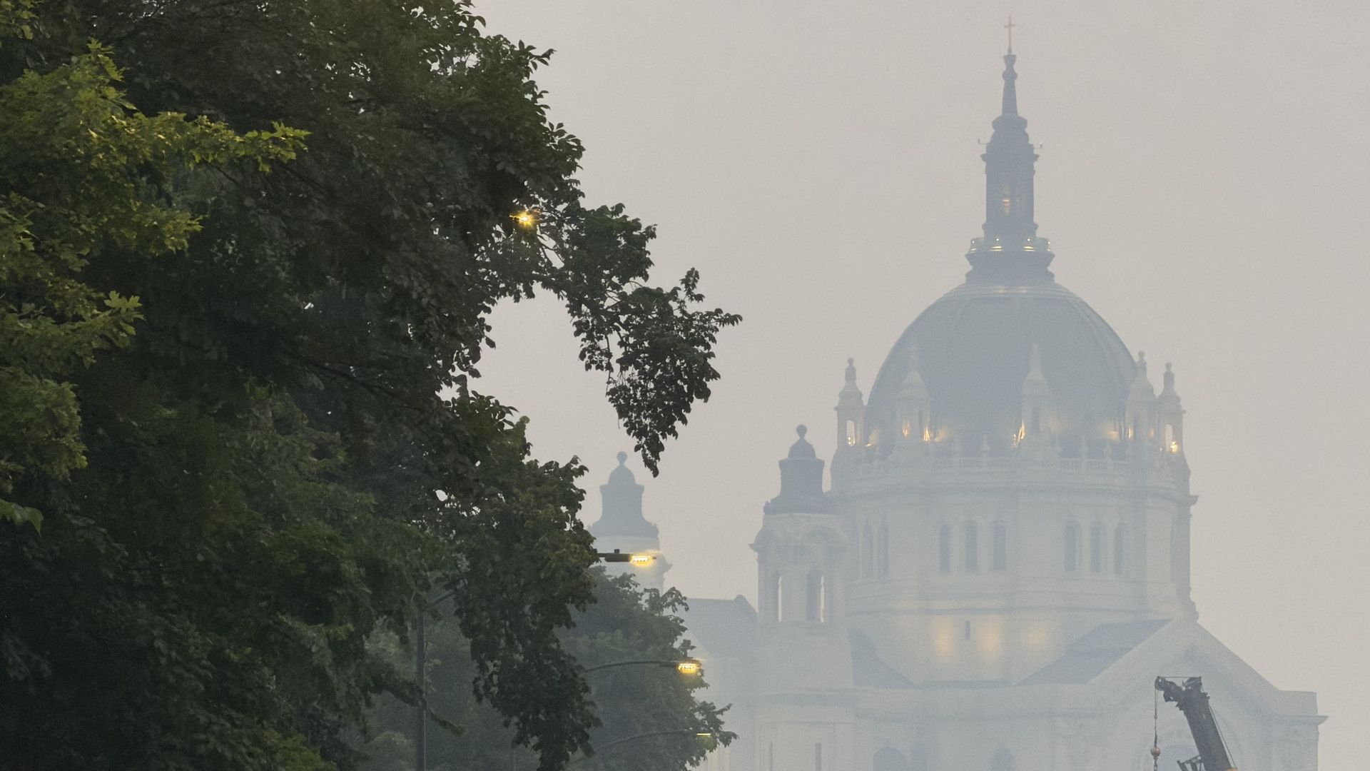 Foggy city scene with a large ornate white domed building on the right, topped by a tall spire with a cross; dark green trees frame the left, a few streetlights glow in the haze.