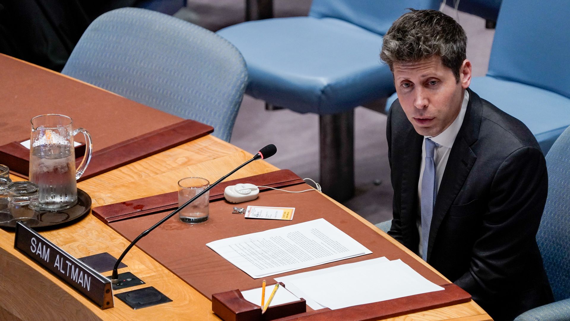Sam Altman speaks into a microphone while sitting behind a curved desk during a UN meeting. He is wearing a dark suit, a white collared shirt and a light blue tie.