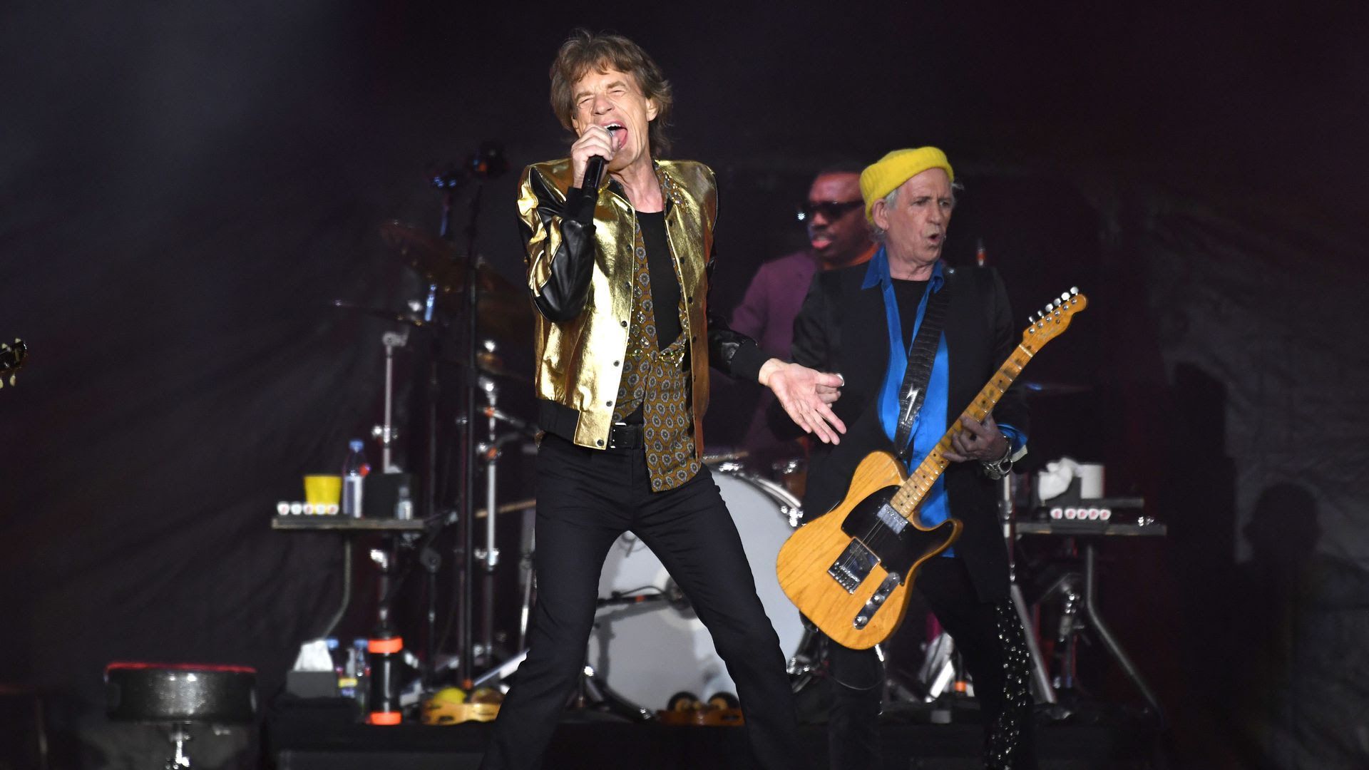 Mick Jagger sings on stage, wearing a gold bomber jacket.