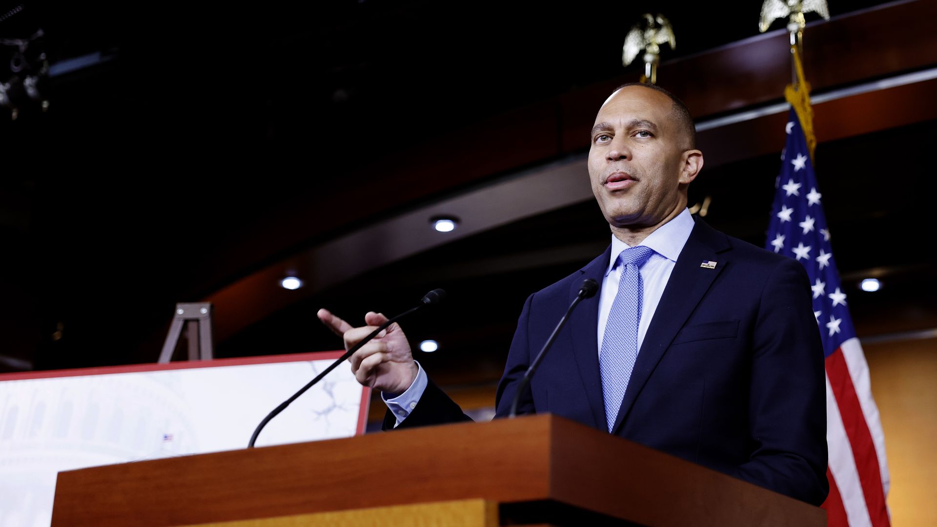 Hakeem Jeffries, wearing a blue suit and speaking at a wooden podium in front of American flags.