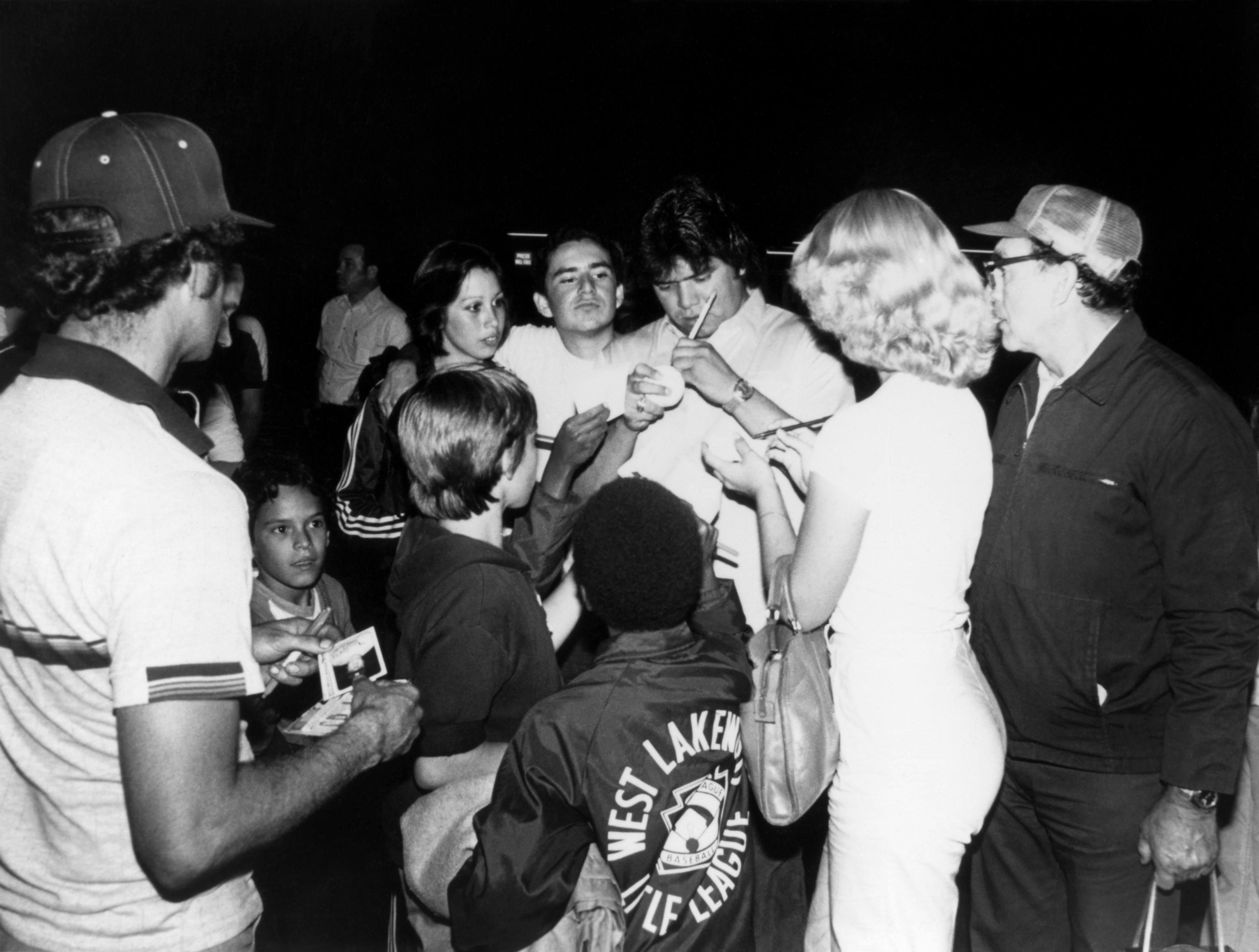 Fernando Valenuzela, shown in black and white, signs an autograph while surrounded by people.