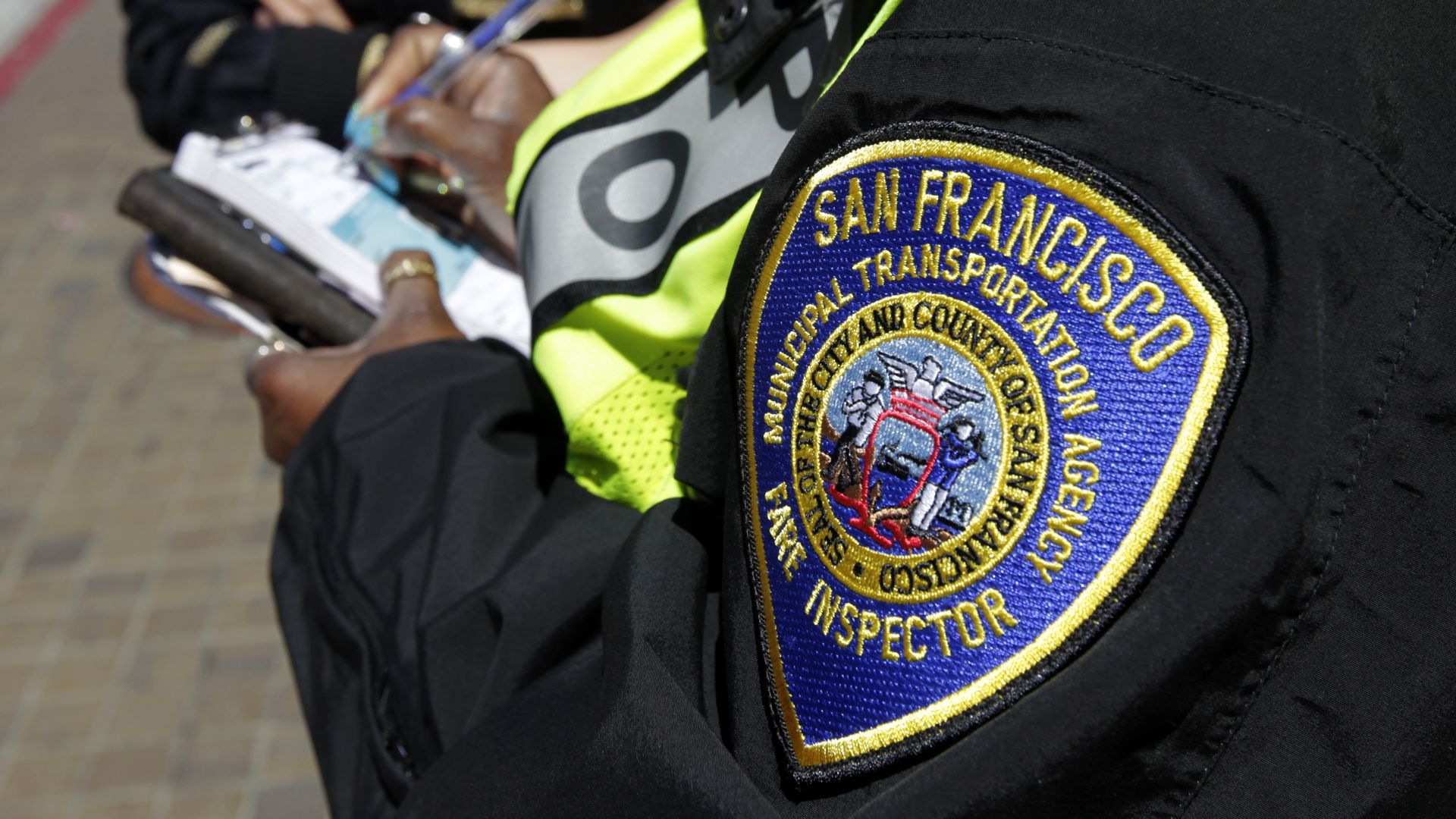 Close-up of a black jacket patch reading "San Francisco Municipal Transportation Agency" in blue and gold; neon-yellow safety vest in the foreground, with a blurred person writing on a clipboard in the background.